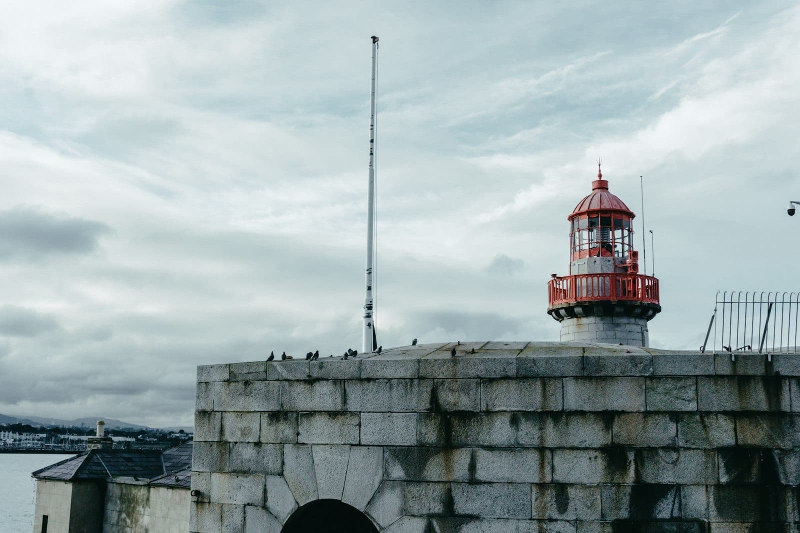 Steinpier und historischer roter Leuchtturm im Hafen von Dún Laoghaire unter bewölktem Himmel, mit Vögeln auf der verwitterten Mauer.