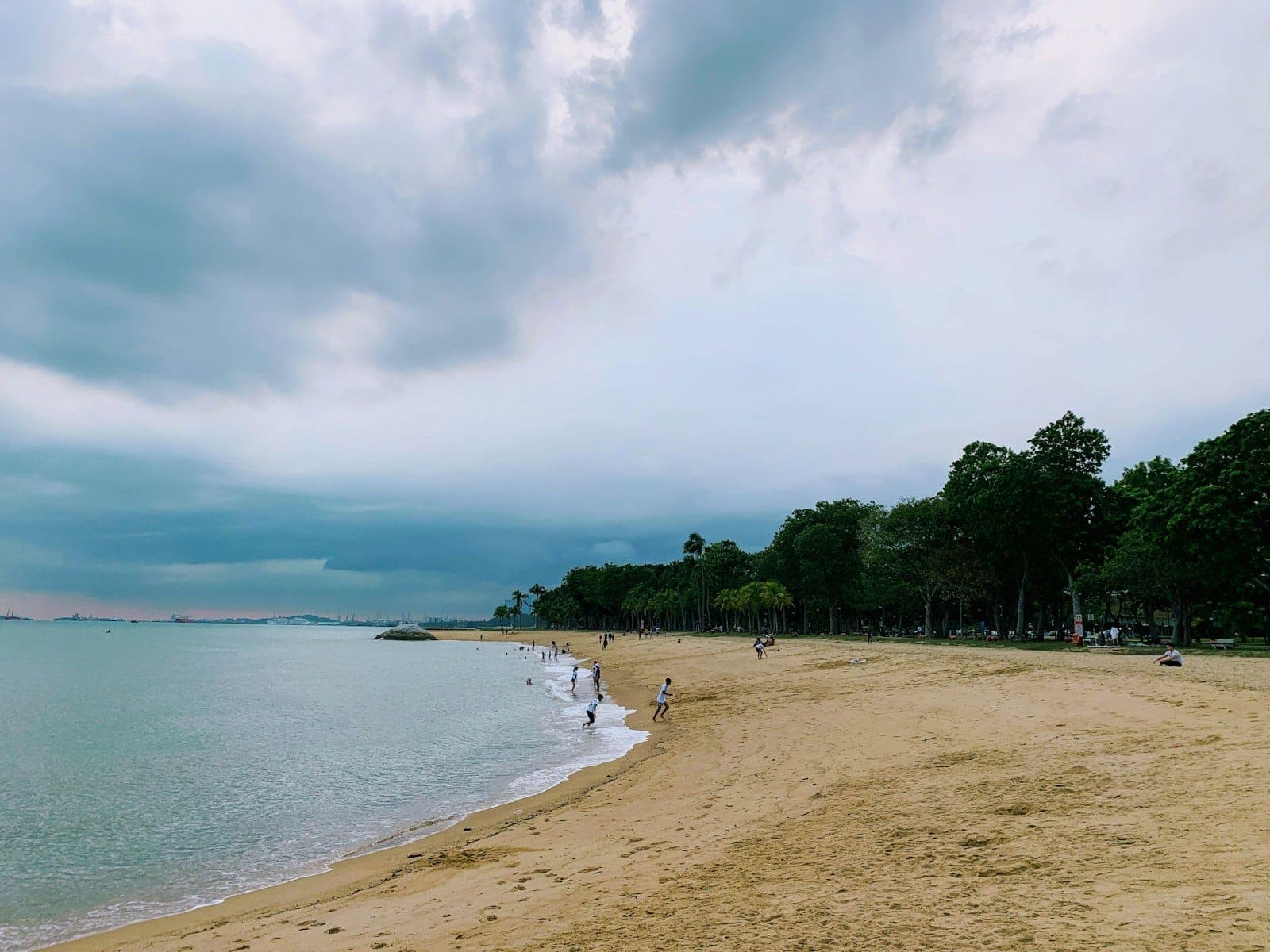 Wide sandy beach with gentle waves, scattered people walking along the shoreline, and lush green trees under a cloudy sky at East Coast Park, Singapore.