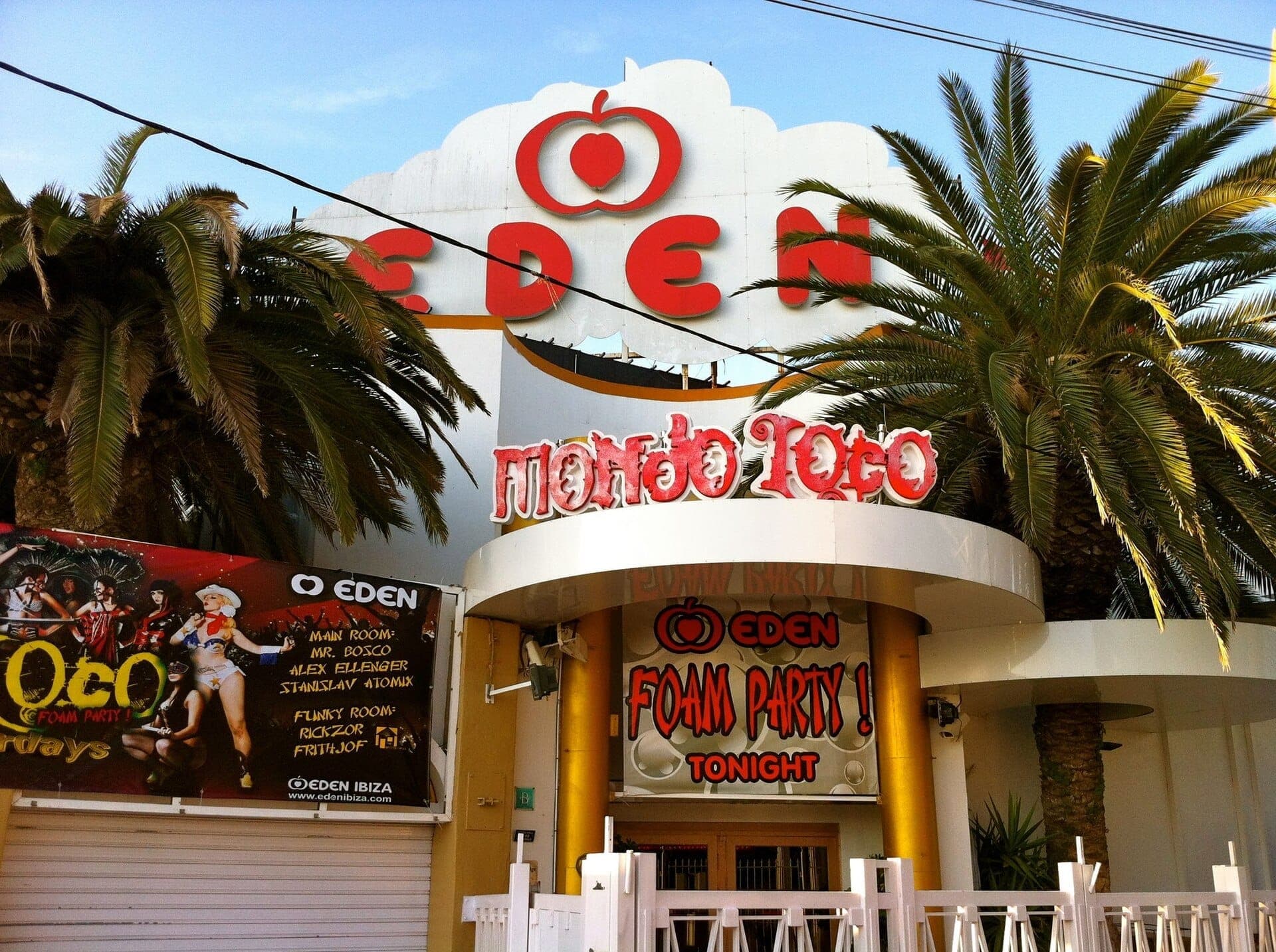 Eden Ibiza superclub entrance with bold red signage, palm trees, foam party banner, and white curved architecture under a clear blue sky.