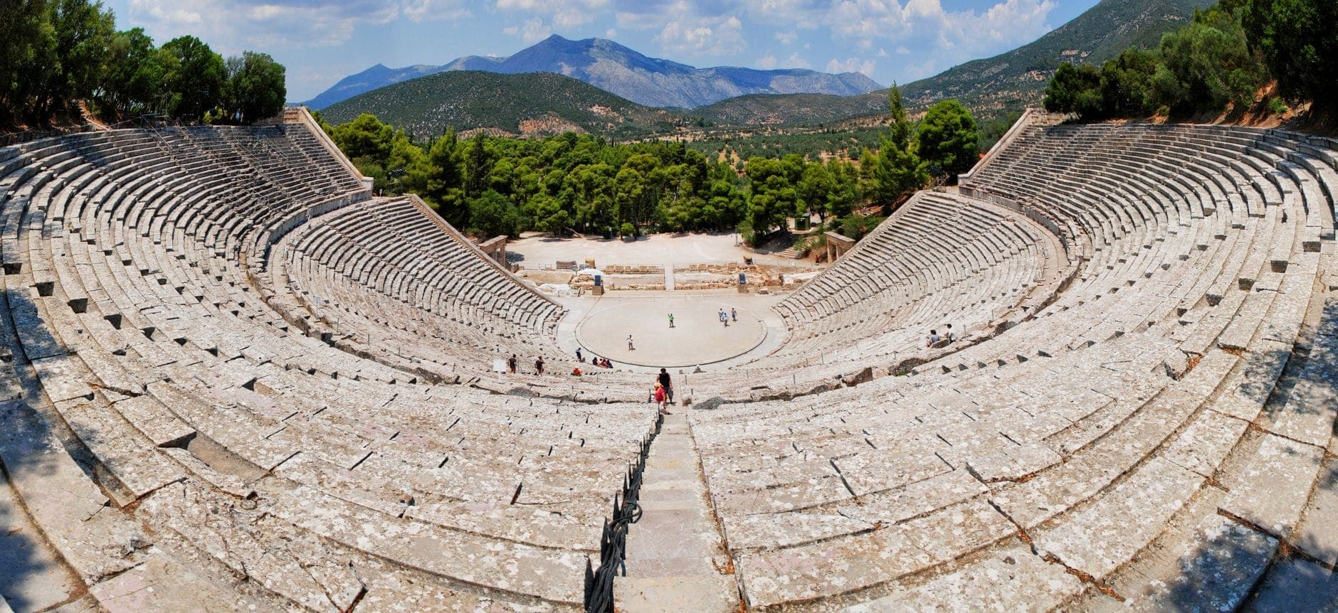 Wide panoramic view of the Ancient Theatre of Epidaurus with sweeping stone seats, surrounded by lush green trees and distant mountains under a blue sky.
