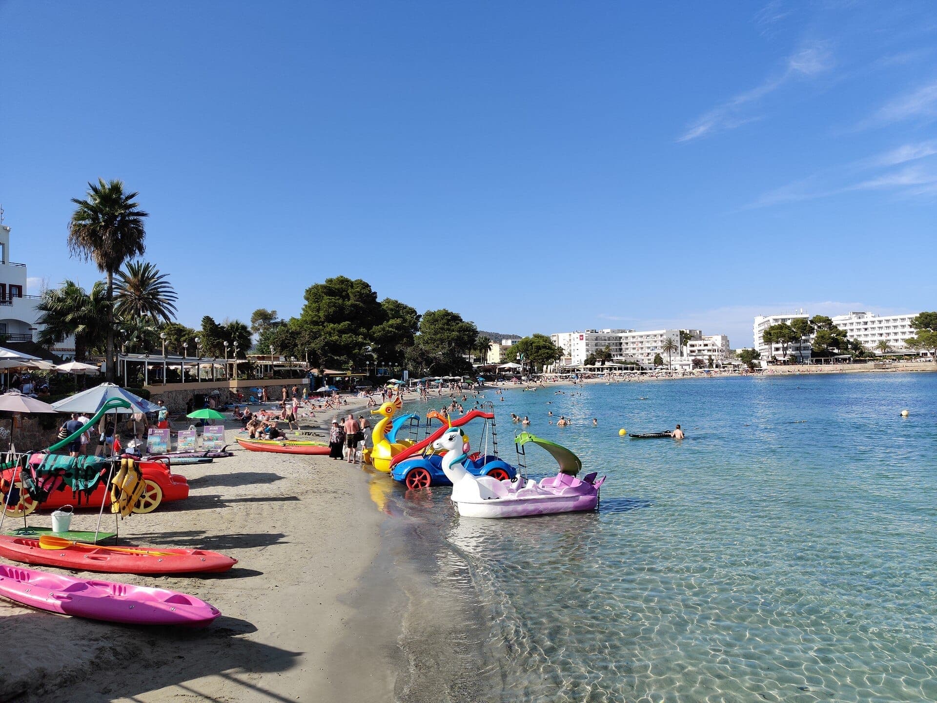 Colorful kayaks and pedal boats line the sandy shore of Es Canar Beach, with sunbathers and swimmers enjoying the clear Mediterranean water.