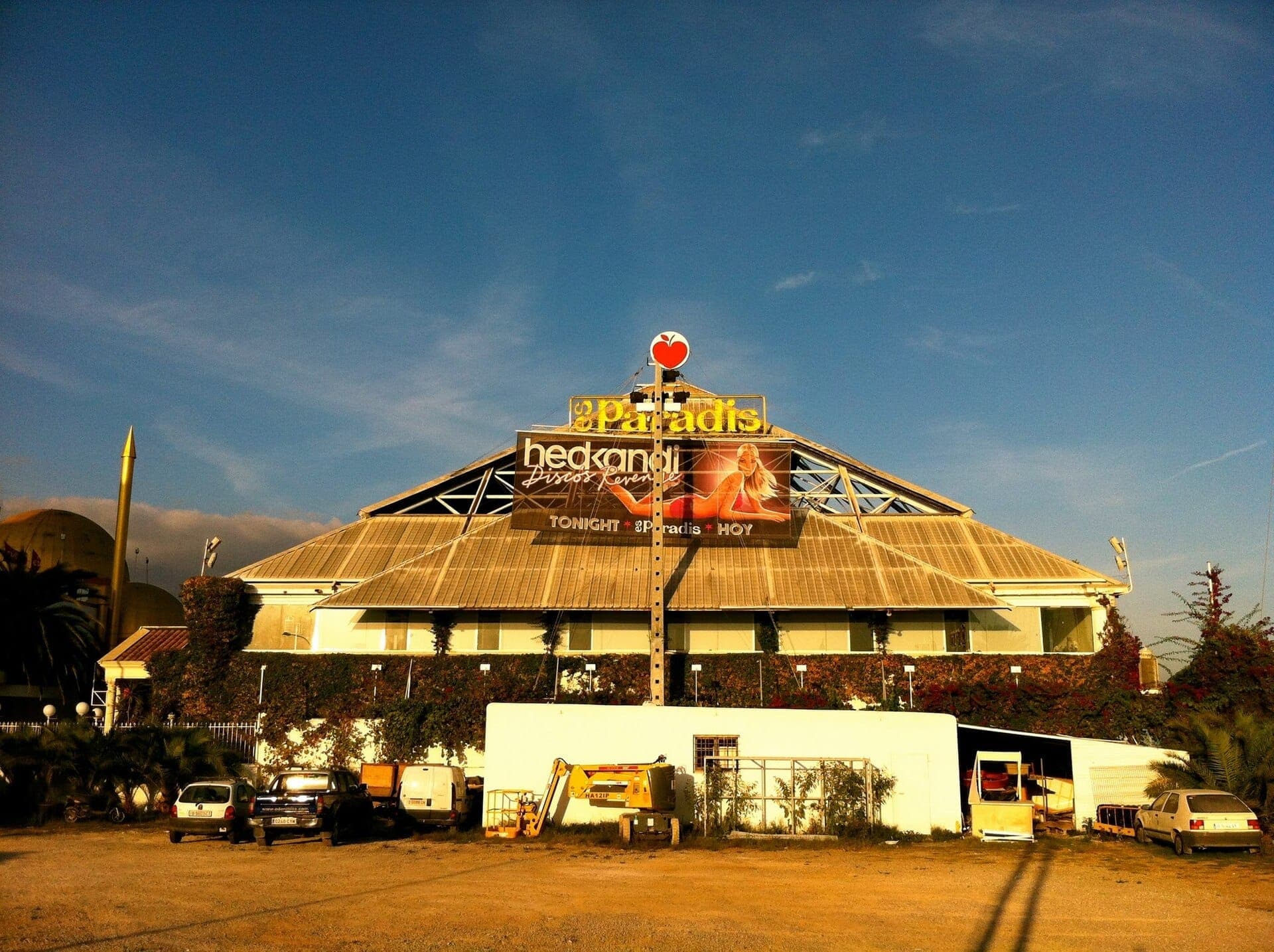 Exterior view of Es Paradis nightclub in Ibiza, featuring its iconic glass pyramid roof, large event banners, and parked vehicles under clear blue skies.