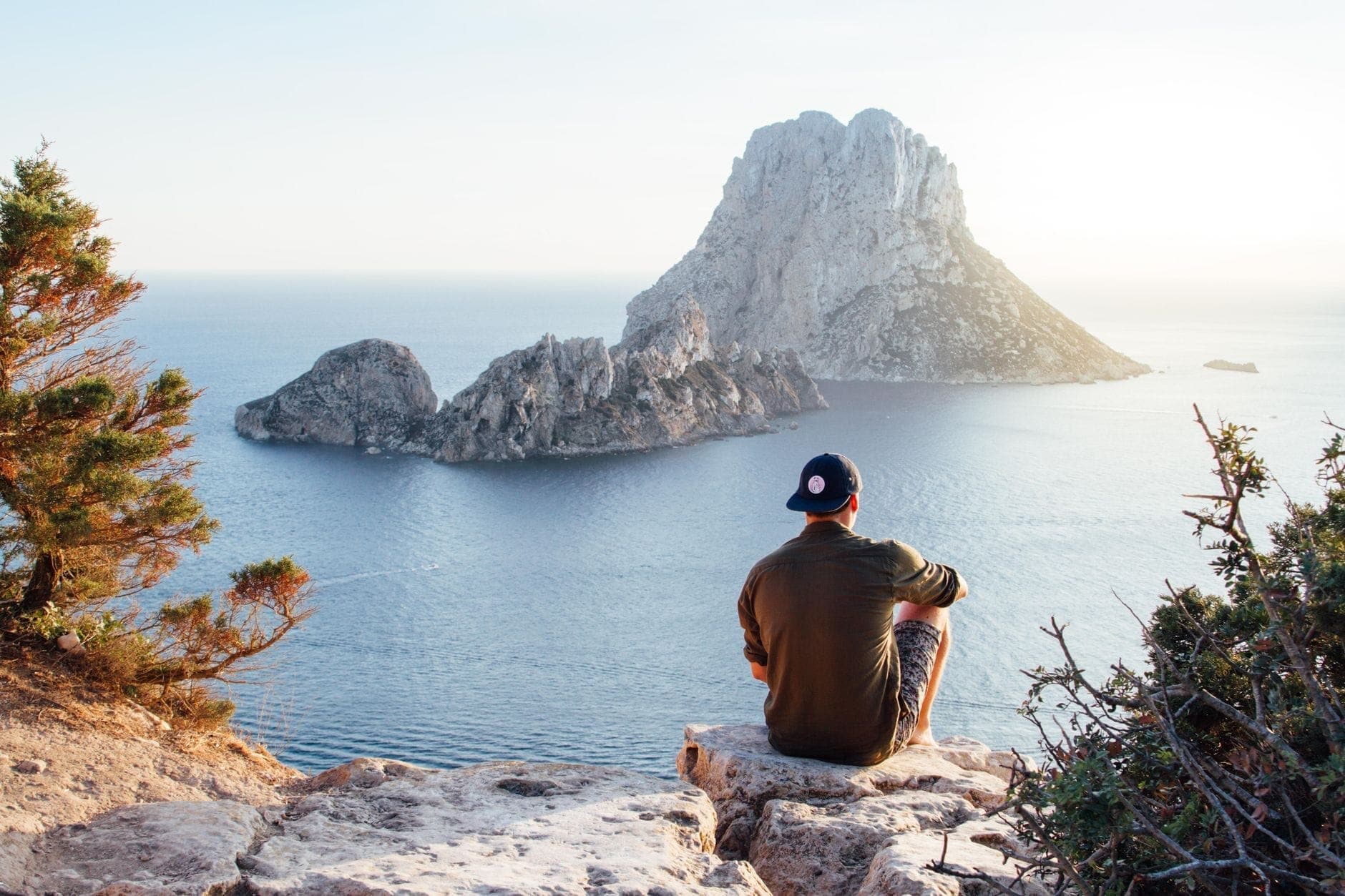A person sits on a rocky cliff overlooking the blue sea and the dramatic limestone island of Es Vedrà near Ibiza.