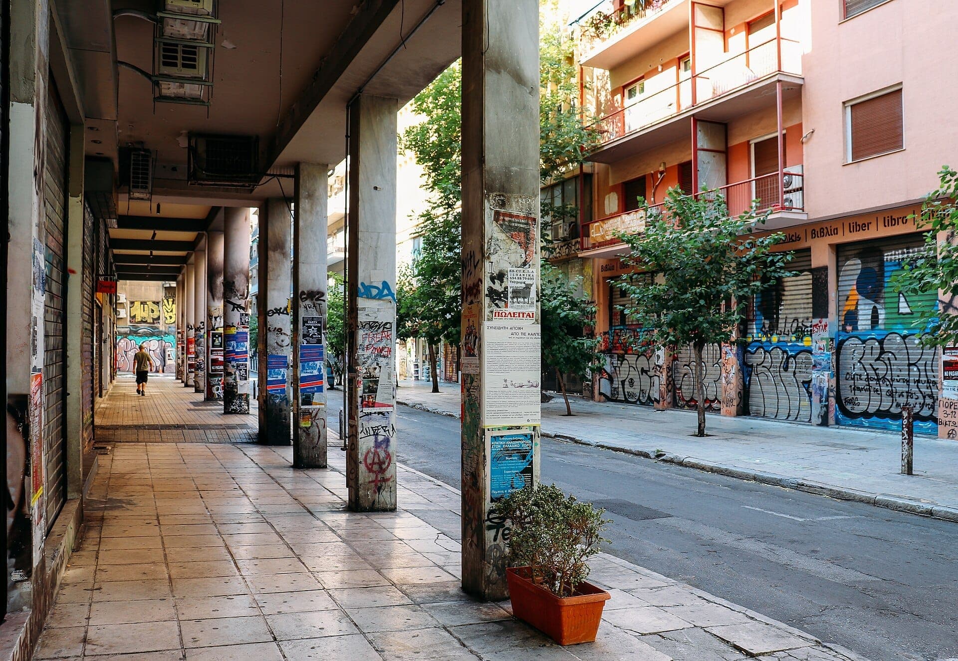 Street scene in Exarchia, Athens, with graffiti-covered buildings, concrete columns plastered with posters, and a lone pedestrian walking under the arcade.