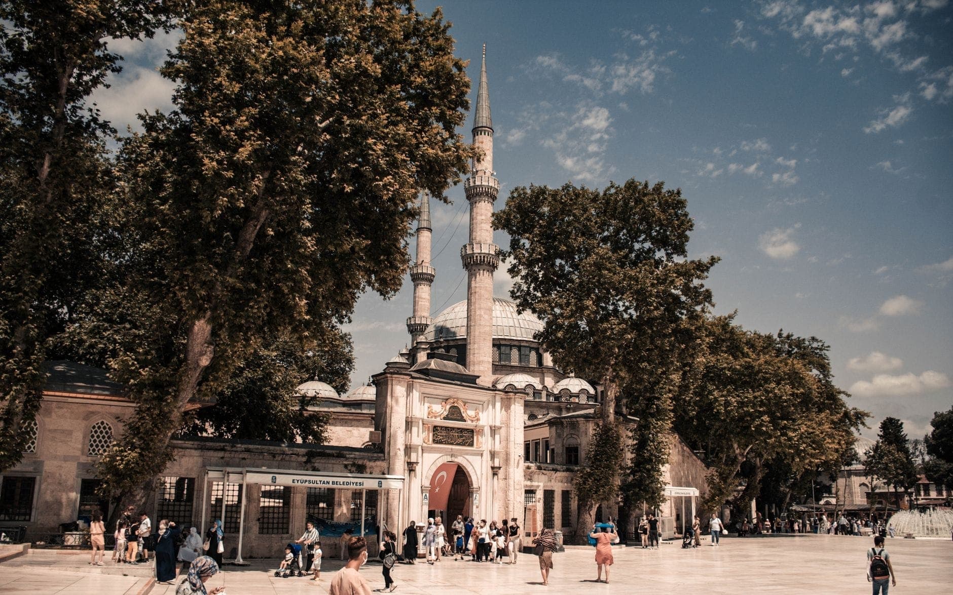 Die Eyüp Sultan Moschee mit ihrem großen Eingangsportal und den Minaretten, umgeben von hohen Bäumen und einem belebten Platz voller Menschen unter hellem Himmel.
