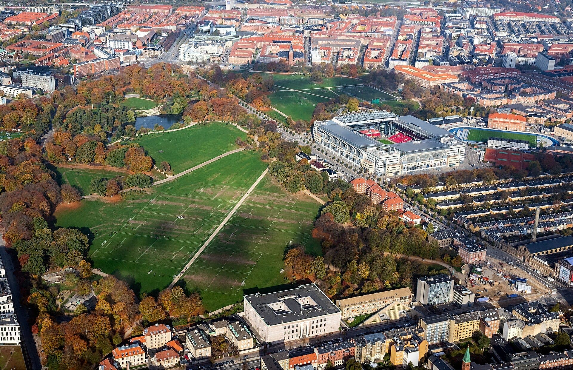 Luftaufnahme des Fælledparken in Kopenhagen mit grünen Rasenflächen, baumgesäumten Wegen, Sportplätzen, einem Teich und einem großen modernen Stadion in der Nähe.
