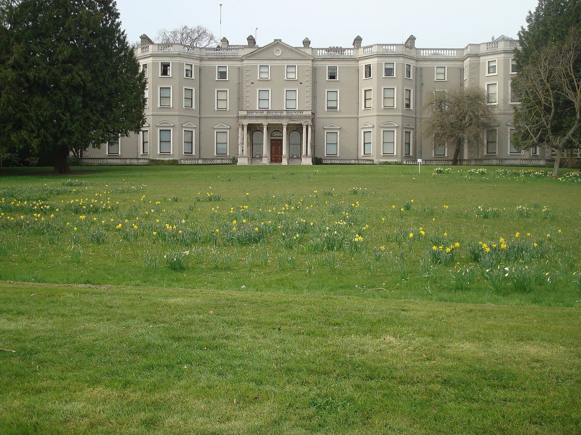 Farmleigh House situada detrás de un extenso césped verde salpicado de narcisos amarillos, enmarcada por árboles maduros en Phoenix Park, Dublín.