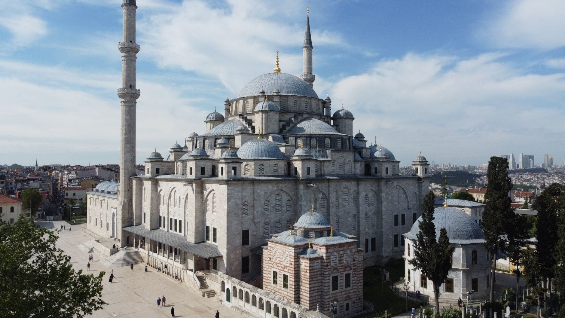 Weitwinkelaufnahme der Fatih-Moschee in Istanbul unter einem teils bewölkten blauen Himmel – mit Kuppeln, Minaretten und dem umliegenden Innenhof, durch den einige Menschen spazieren.