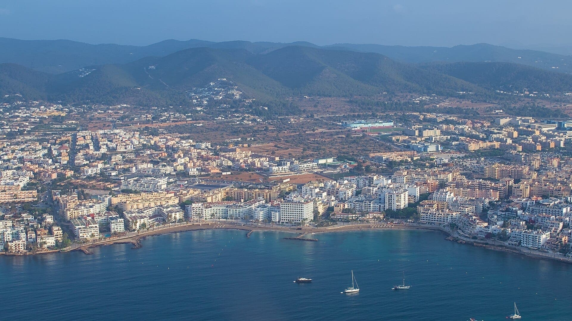 Aerial view of Figueretas Beach in Ibiza Town, with urban buildings, sandy shoreline, calm blue water, boats, and distant green hills.