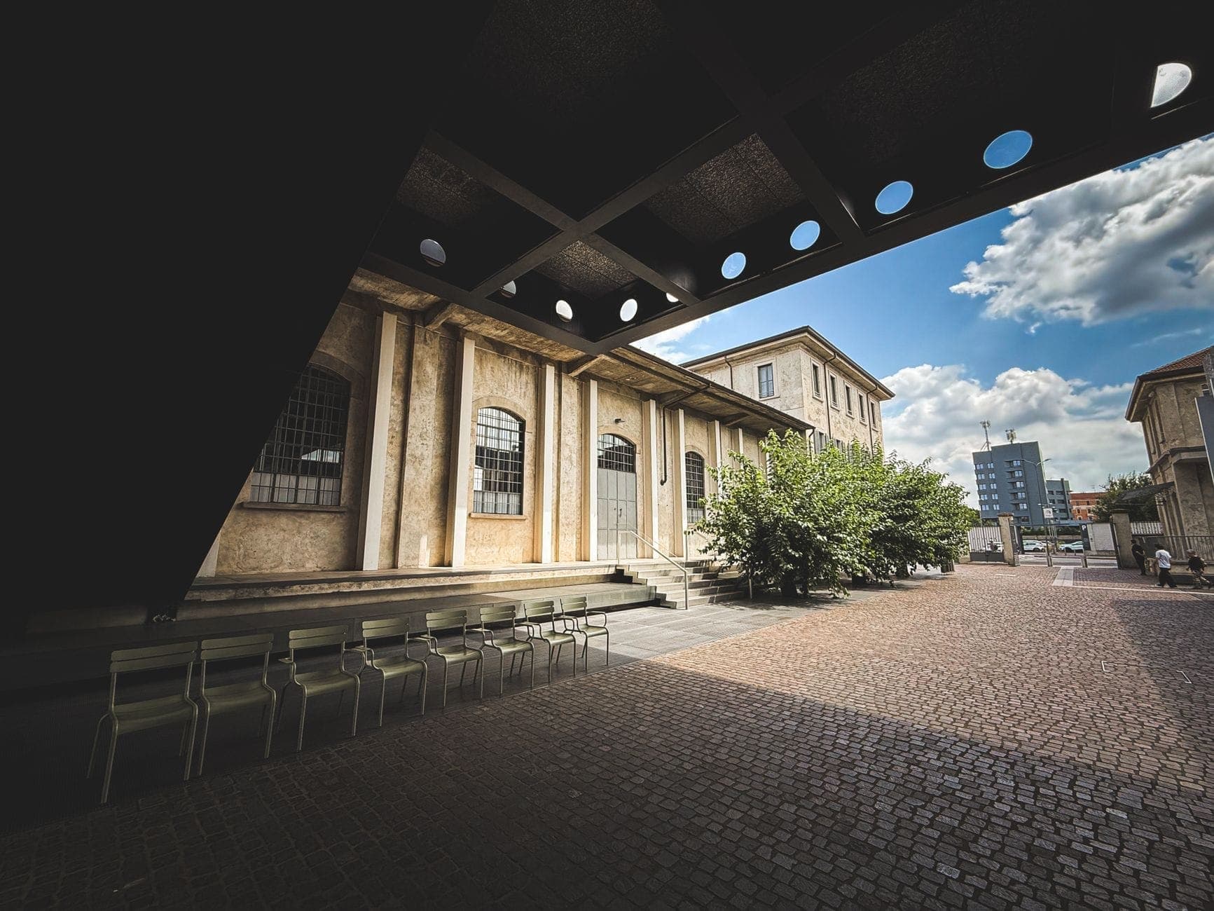 View of Fondazione Prada’s distinctive courtyard with modern industrial architecture, dramatic overhang, rows of chairs, and historic distillery buildings under a bright blue sky.