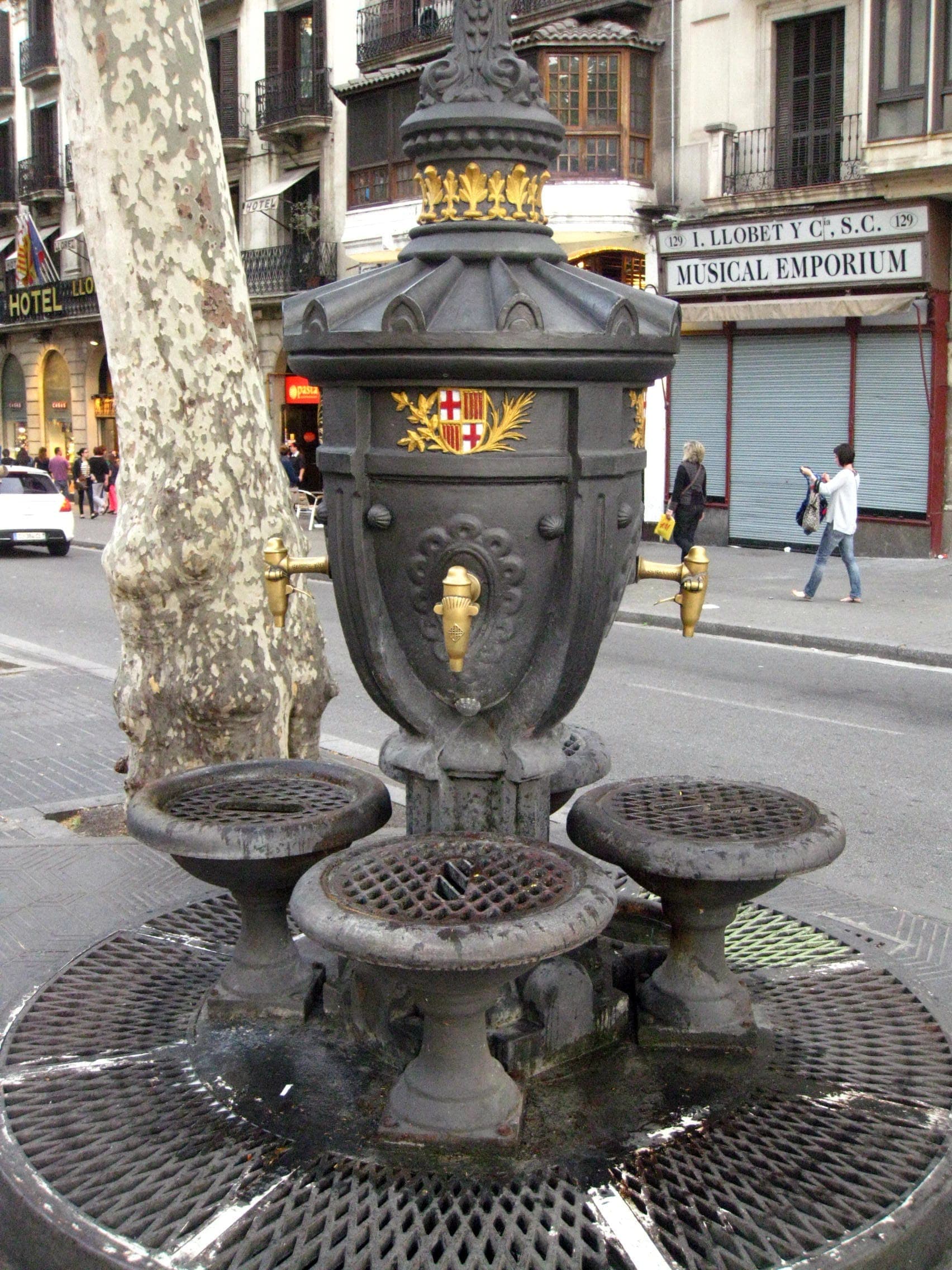 Đài phun nước Font de Canaletes bằng gang đen được trang trí tinh xảo với các chi tiết mạ vàng, đứng bên cạnh một cái cây trên con phố nhộn nhịp ở Barcelona.
