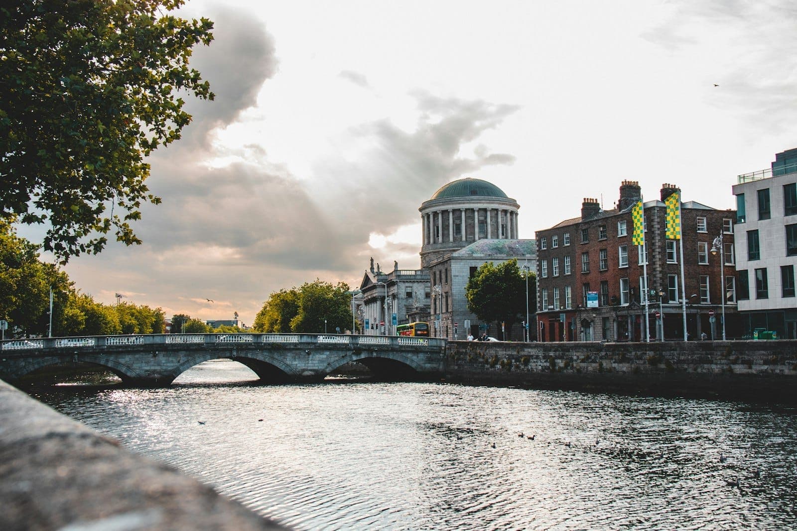 Vista amplia de los Four Courts de Dublín con su cúpula y fachada neoclásica junto al río Liffey y un puente de piedra bajo un cielo nublado y dramático.