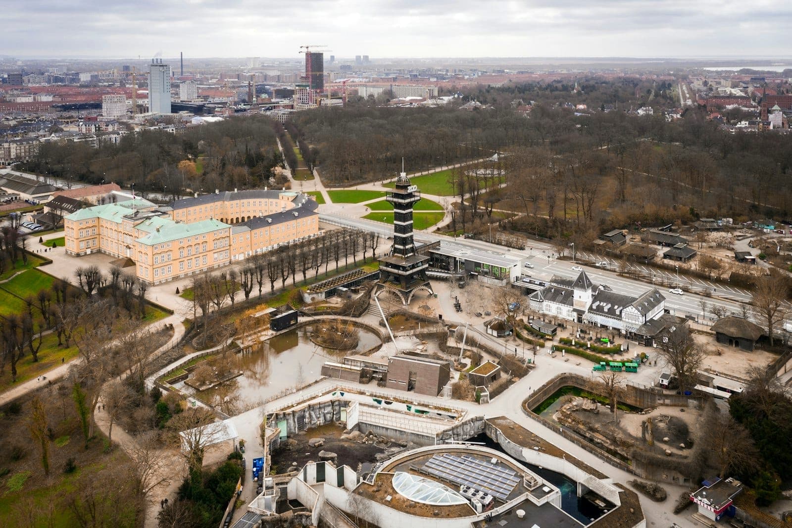 Luftaufnahme von Schloss Frederiksberg und den umliegenden Gärten, mit der Stadtsilhouette im Hintergrund unter einem bewölkten Himmel.