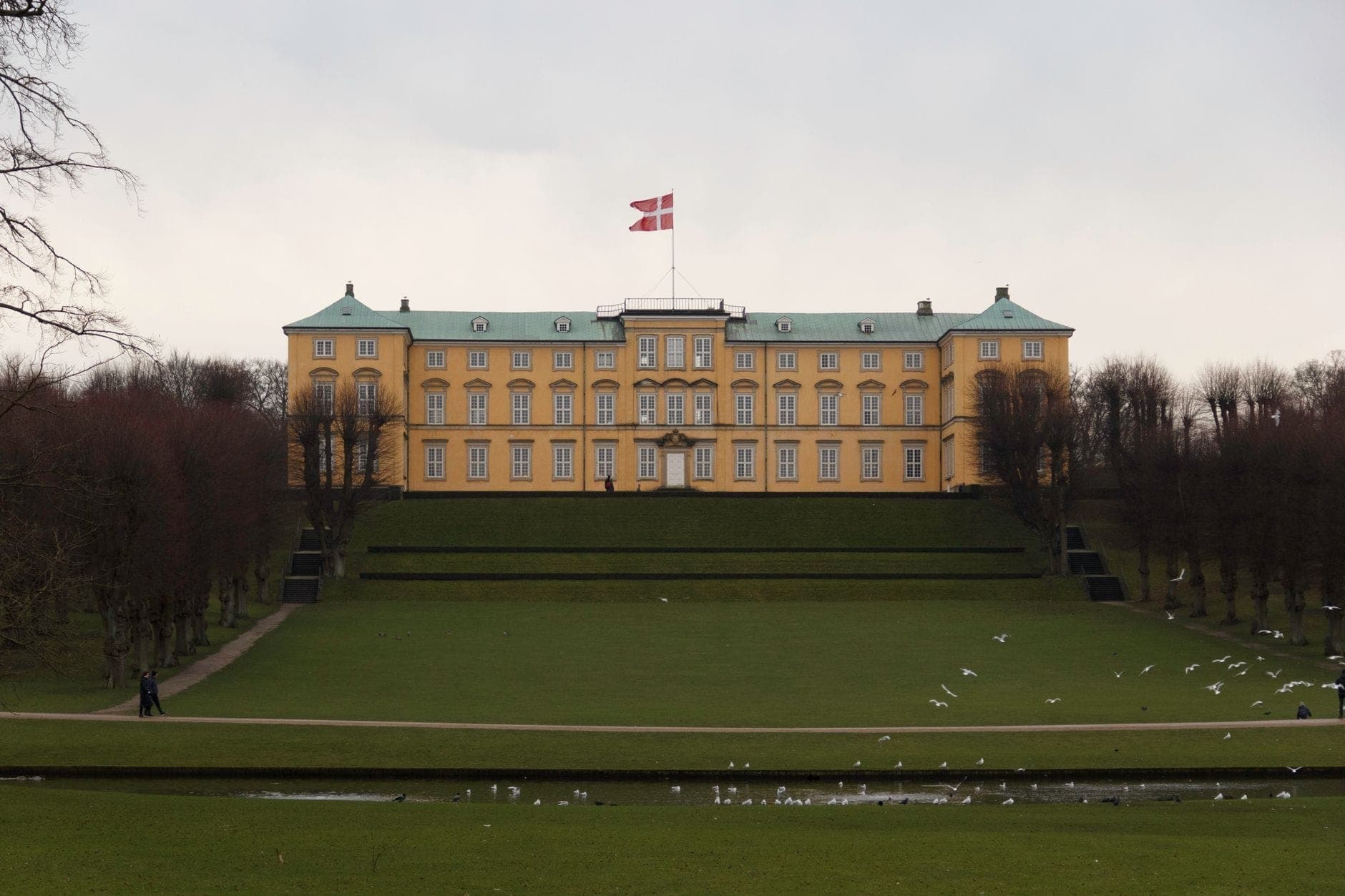 Il Palazzo di Frederiksberg a Copenaghen, un imponente edificio barocco giallo in cima a una collina, con la bandiera danese che sventola e ampi prati paesaggistici in primo piano.