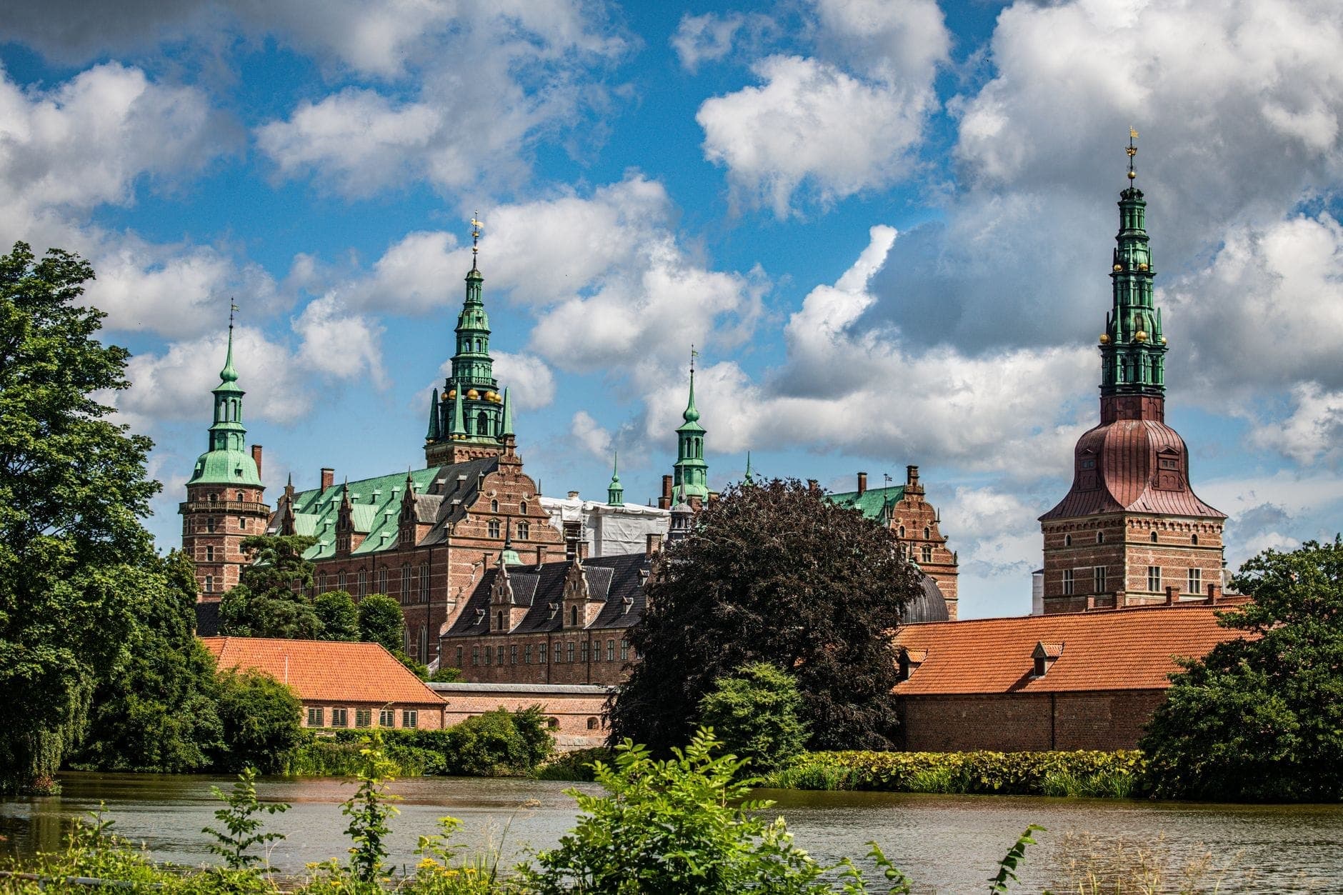 Frederiksborg Castle in Denmark viewed from across a lake, with green copper towers rising above red brick buildings under a bright blue sky and scattered clouds.