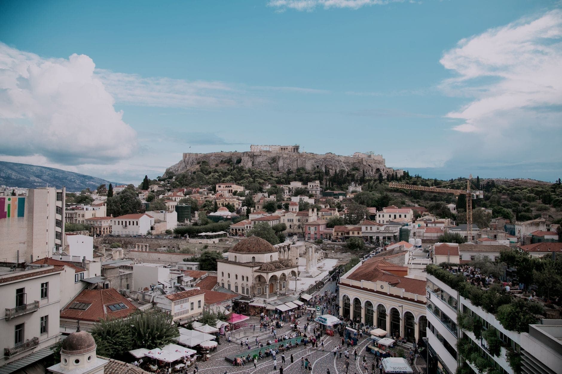 Ein weiter Blick auf Athen mit dem belebten Monastiraki-Platz voller Menschen, Märkten, historischen Gebäuden und der Akropolis im Hintergrund unter einem blauen Himmel.