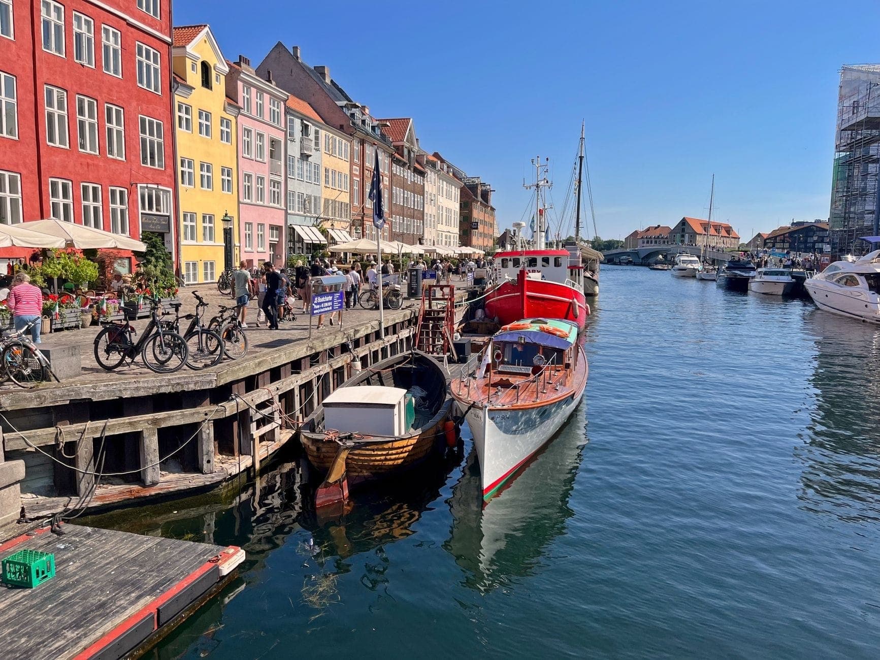 Bunte historische Häuser und belebte Cafés säumen den Nyhavn-Kanal in Kopenhagen, mit festgemachten Booten und Menschen, die einen sonnigen Tag genießen.