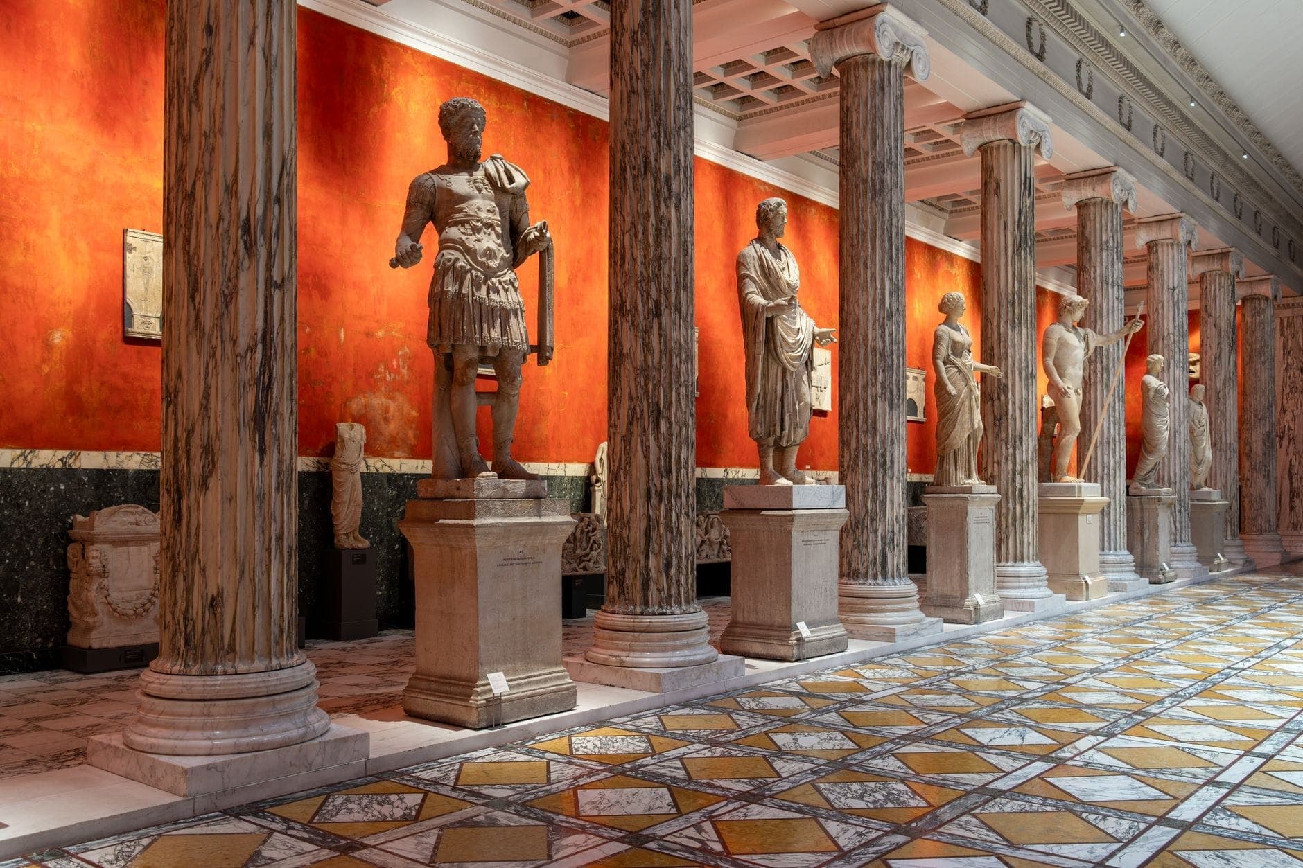 Bulwagan ng isang museo ng sining na may mataas na mga haligi ng marmol at mga estatuwang estilo Romano laban sa isang kapansin-pansing pulang dingding, malamang sa Copenhagen.