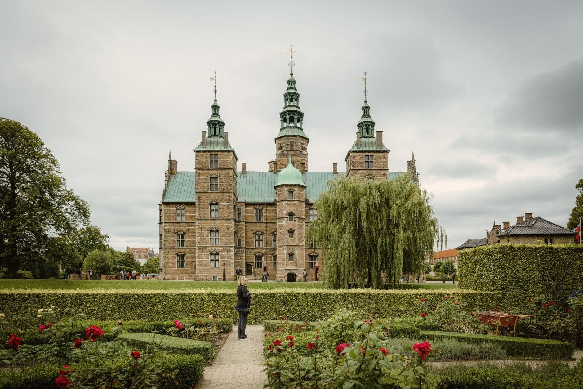 Simetrikal na pagtingin sa Rosenborg Castle na may mga pormal na hardin sa harap, namumukadkad na mga bulaklak, isang taong naglalakad, at malusog na mga berdeng bakod sa Copenhagen.