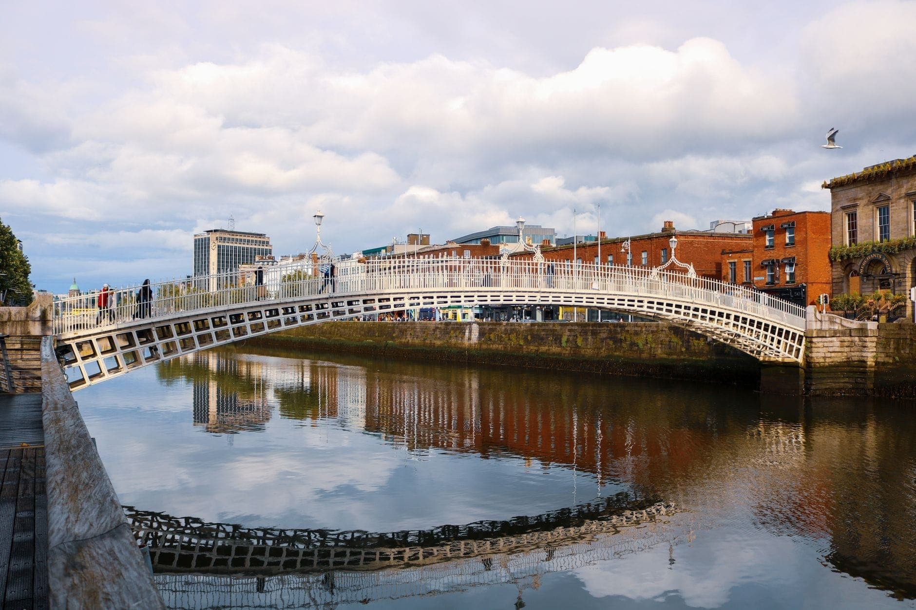 Vista diurna del Ha'penny Bridge cruzando el río Liffey en Dublín, con peatones, edificios de ladrillo rojo y reflejos en el agua.