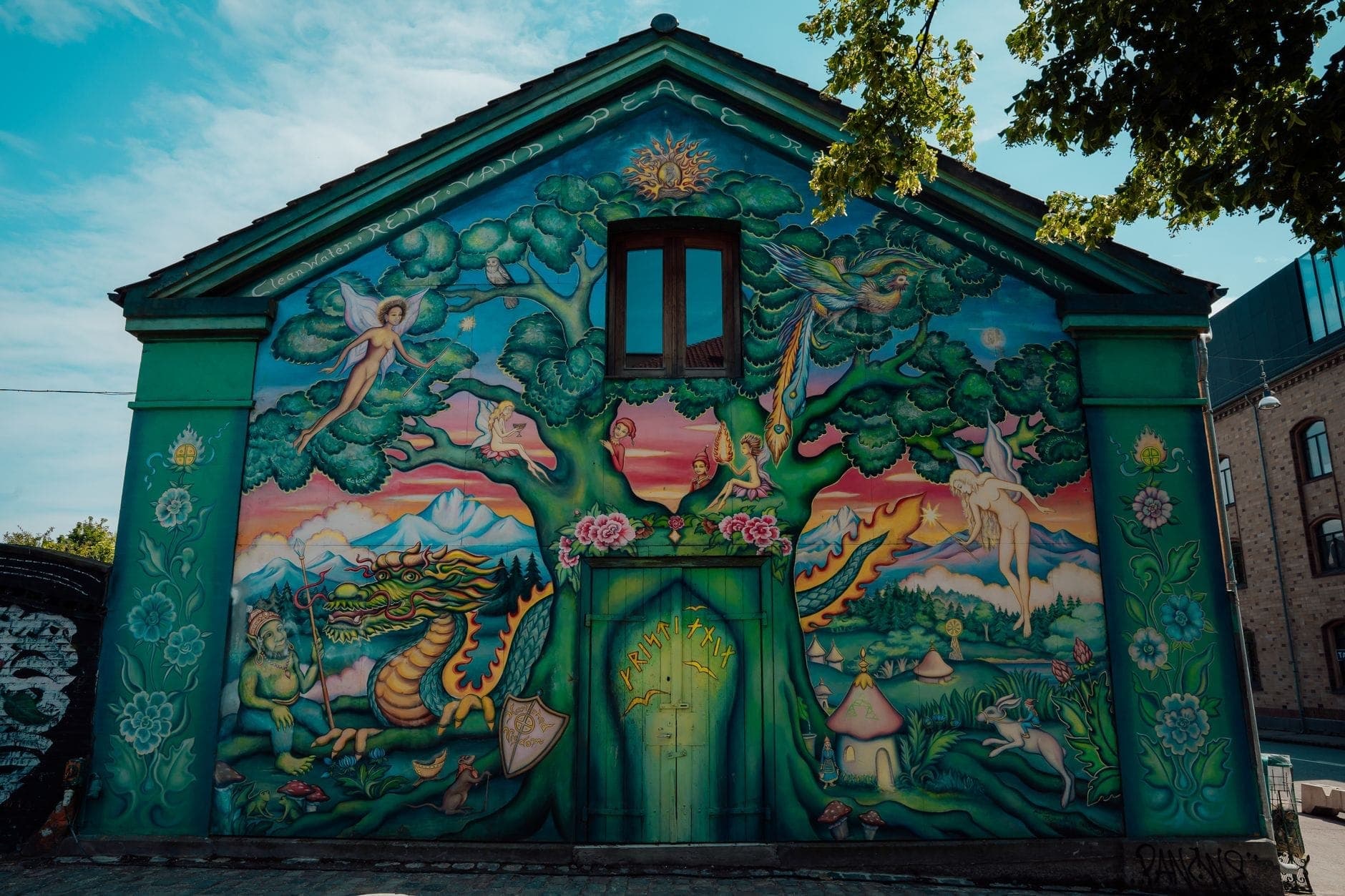 Buntes Wandgemälde an der Fassade eines Gebäudes in Freetown Christiania mit einer farbenfrohen Fantasieszene — Baum, Drache, Fee und mystische Elemente vor blauem Himmel.
