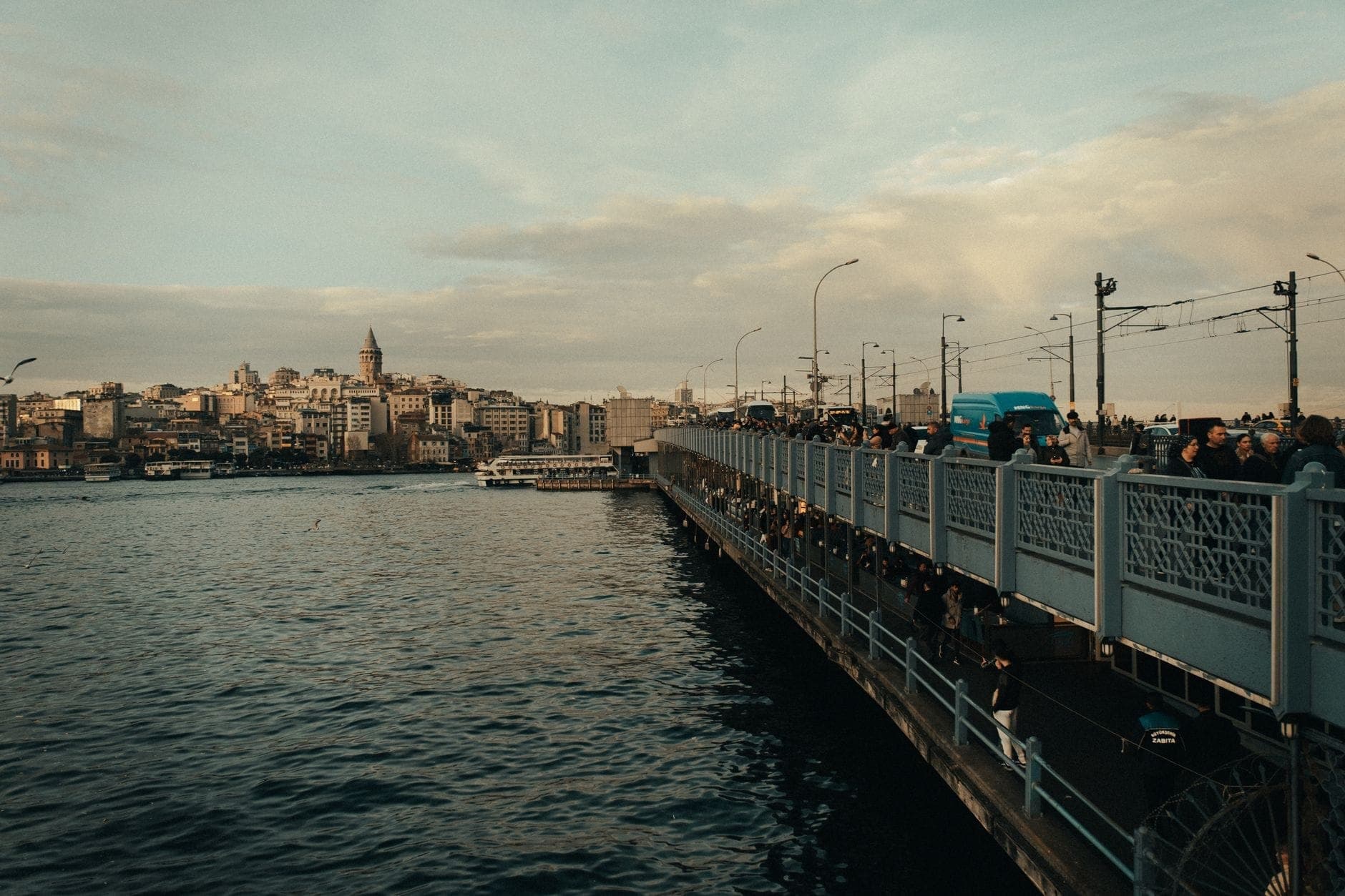Die Galata-Brücke in Istanbul in der Abenddämmerung mit Menschen beim Spazieren, Angeln und Fahrzeugen, die die Brücke überqueren – im Hintergrund die Stadtsilhouette mit dem Galata-Turm.