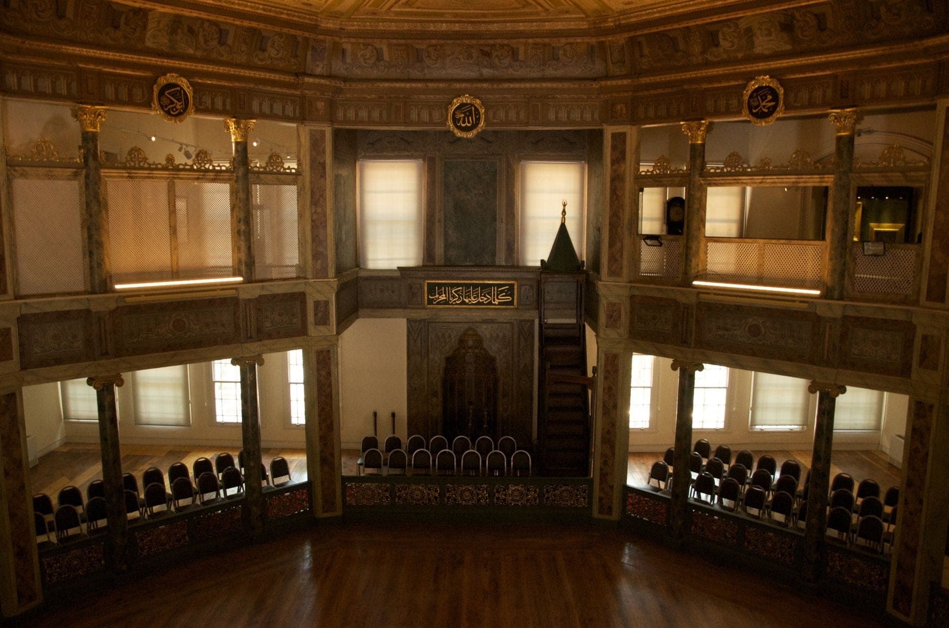 Innenraum des Galata-Mevlevi-Museums mit Holzböden, verziertem Balkon, dekorativer Kalligraphie und Reihen leerer Stühle im sanften Fensterlicht.