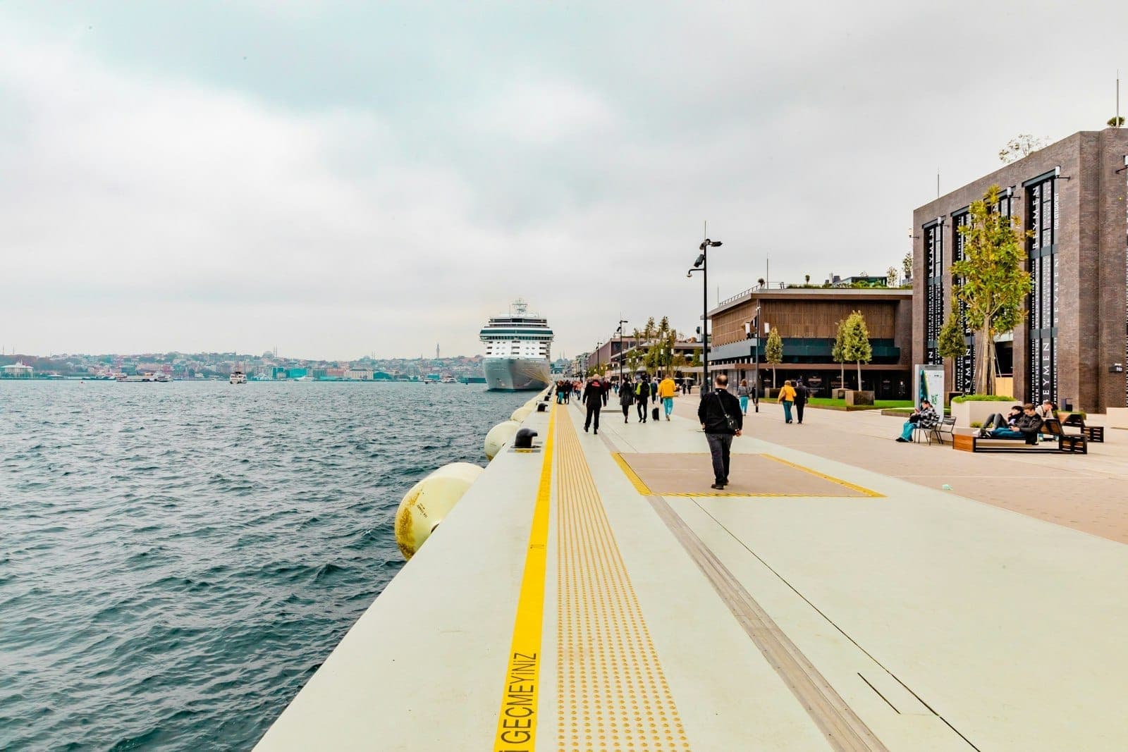 Galataport Istanbul Uferpromenade mit Spaziergängern, modernen Gebäuden auf der rechten Seite und einem am Bosporus angedockten Kreuzfahrtschiff an einem bewölkten Tag.