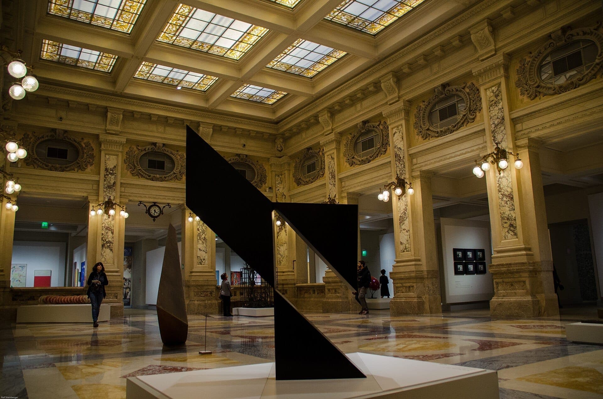 Visitors explore the grand marble hall and modern sculptures at Gallerie d’Italia – Piazza Scala in Milan, under ornate stained-glass ceilings.