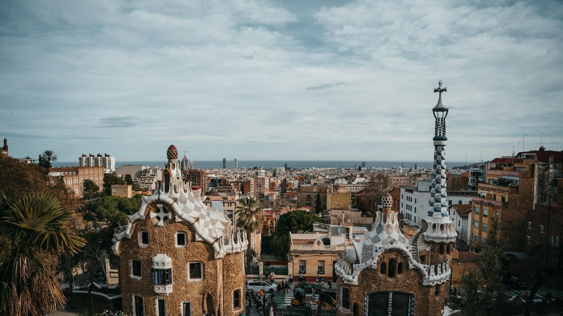 Góc nhìn rộng trên Park Güell với những ngôi nhà và tháp đỉnh đầy màu sắc kỳ ảo, phía sau là toàn cảnh thành phố Barcelona và biển Địa Trung Hải dưới bầu trời có mây.
