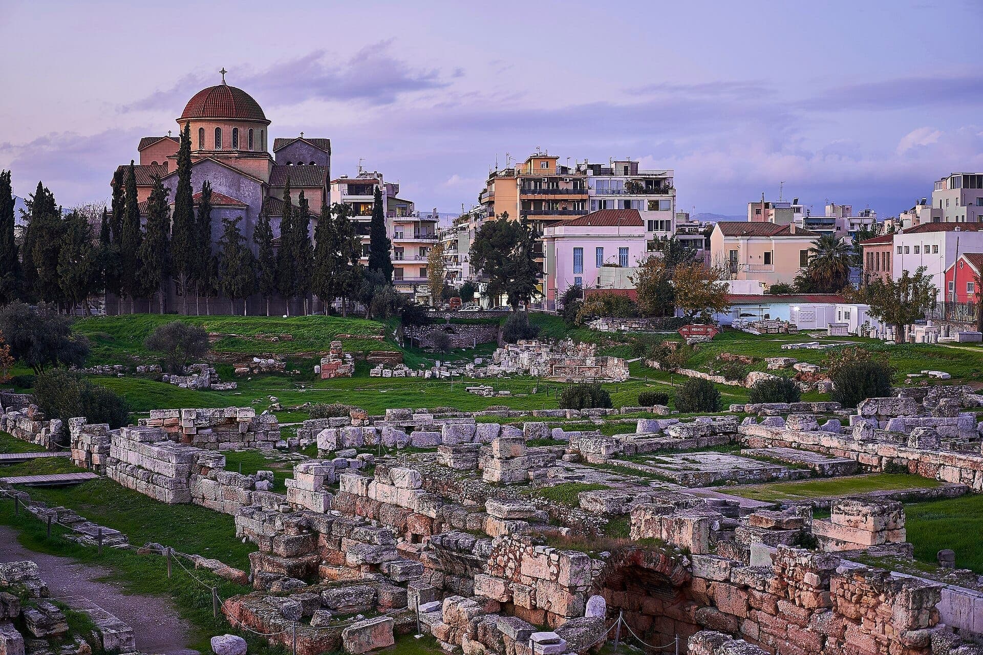Ancient stone ruins of Kerameikos cemetery with a domed church and colorful urban buildings in the background under a soft evening sky.