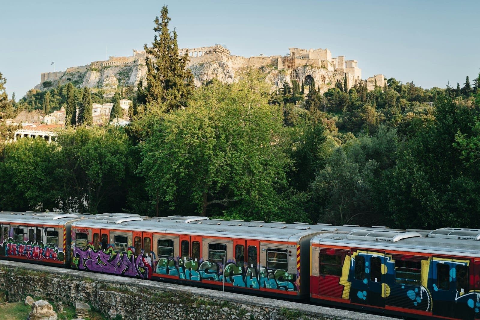 Ein bunter, mit Graffiti besprühter Metrozug fährt vor üppigem Grün entlang – im Hintergrund thront die Akropolis über der Stadt, Symbol für Athens öffentlichen Nahverkehr und seine Wahrzeichen.