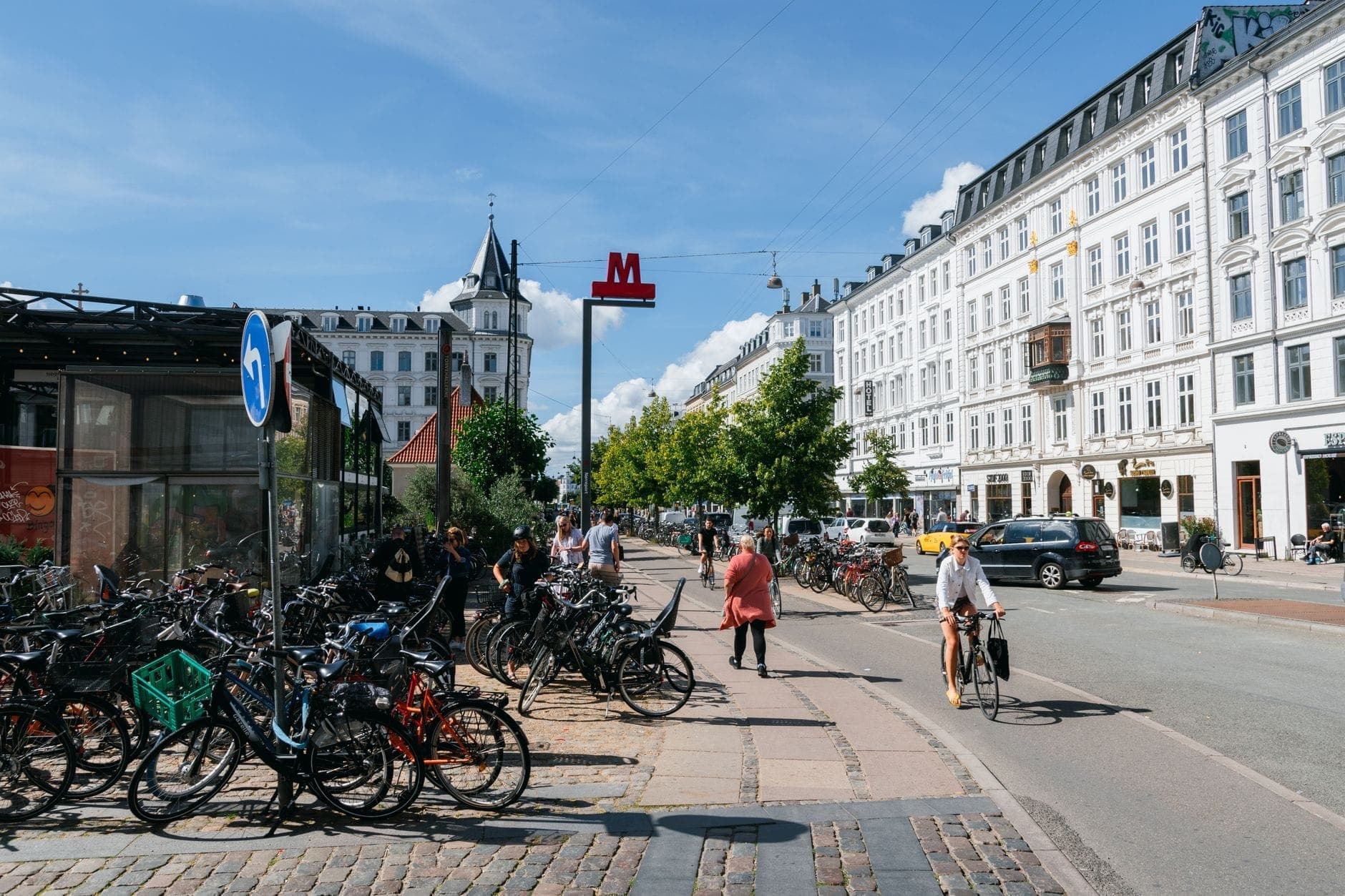 Fahrräder parken entlang einer Stadtstraße; Menschen radeln und laufen an einem U-Bahn-Eingang im Herzen Kopenhagens an einem sonnigen Tag vorbei.