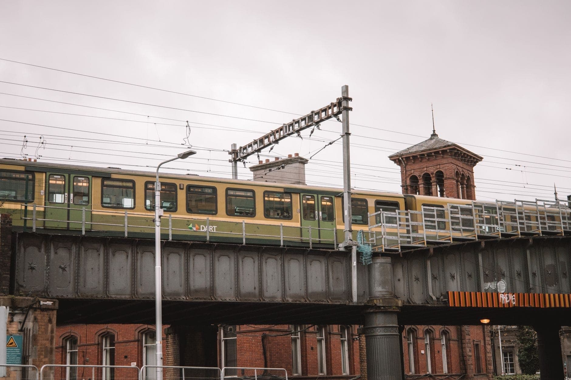 Ein DART-Zug überquert eine erhöhte Eisenbahnbrücke in Dublin, mit historischen Backsteingebäuden und einem Uhrturm im Hintergrund.