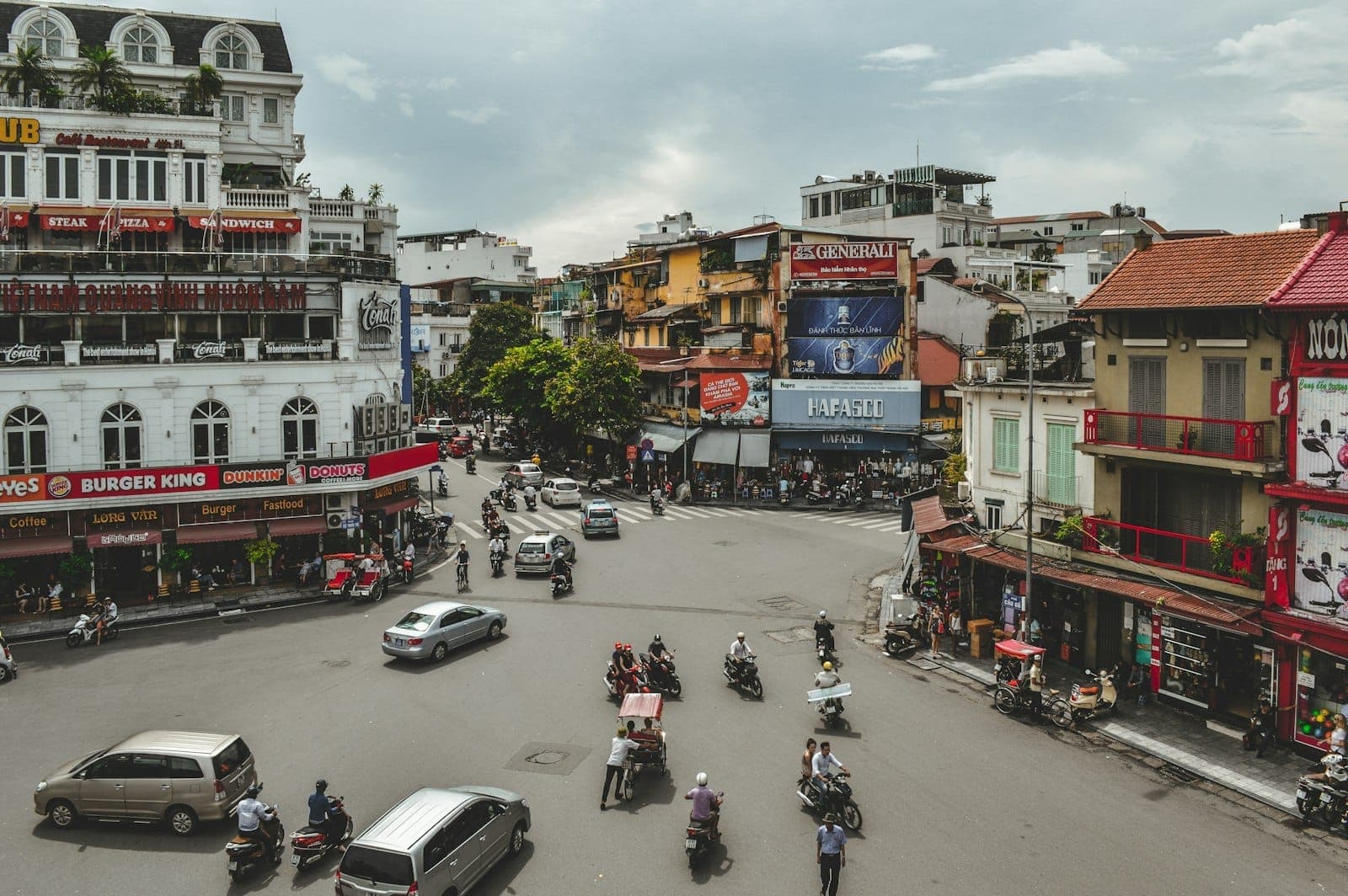 Ngã tư đông đúc ở Hà Nội với ô tô, xe máy, xích lô và người đi bộ chen chúc giữa những tòa nhà và cửa hàng đầy màu sắc trong một ngày nhiều mây.