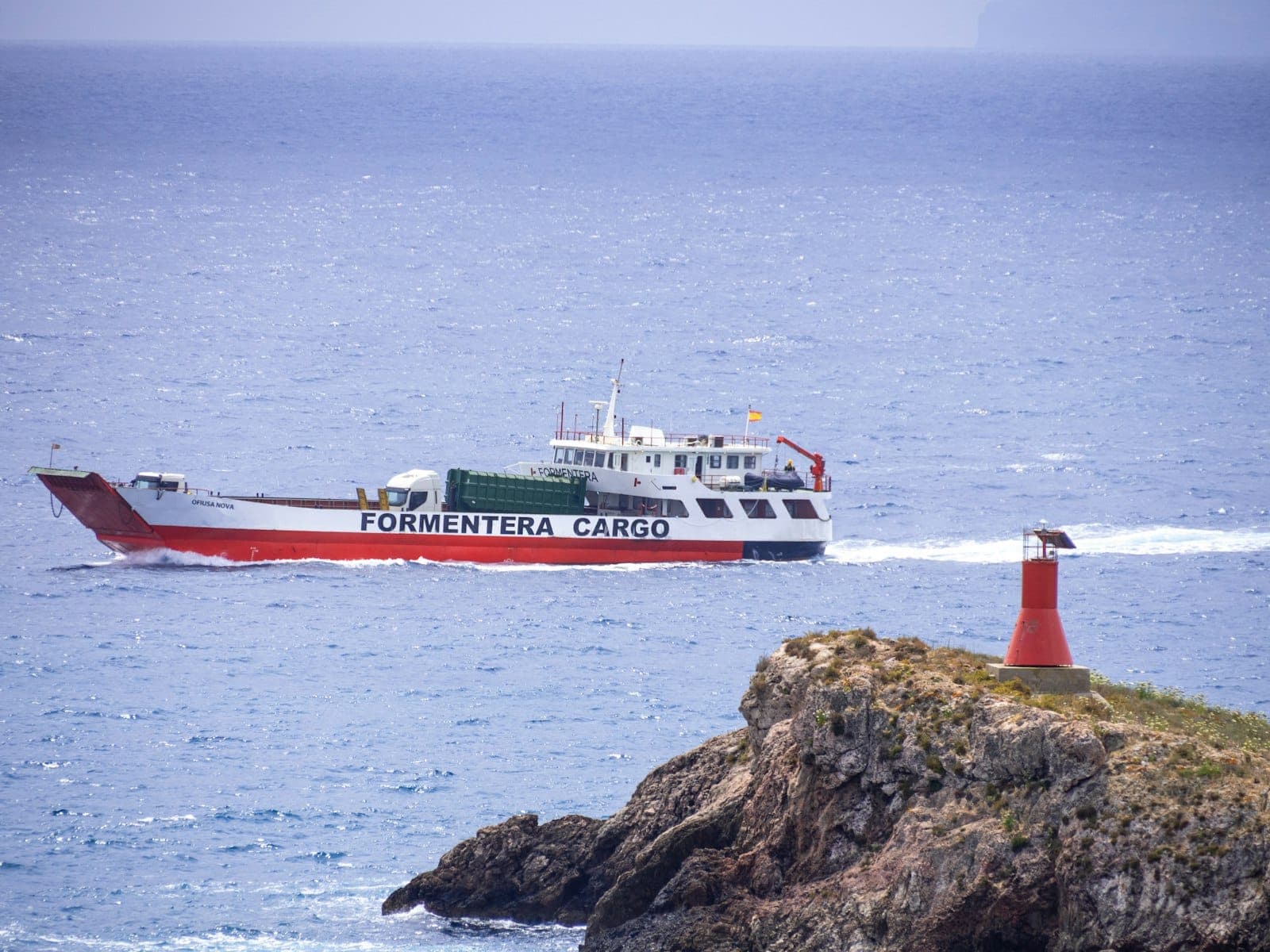 A ferry labeled 'Formentera Cargo' sails past a rocky headland with a red navigation beacon on the coast near Ibiza.