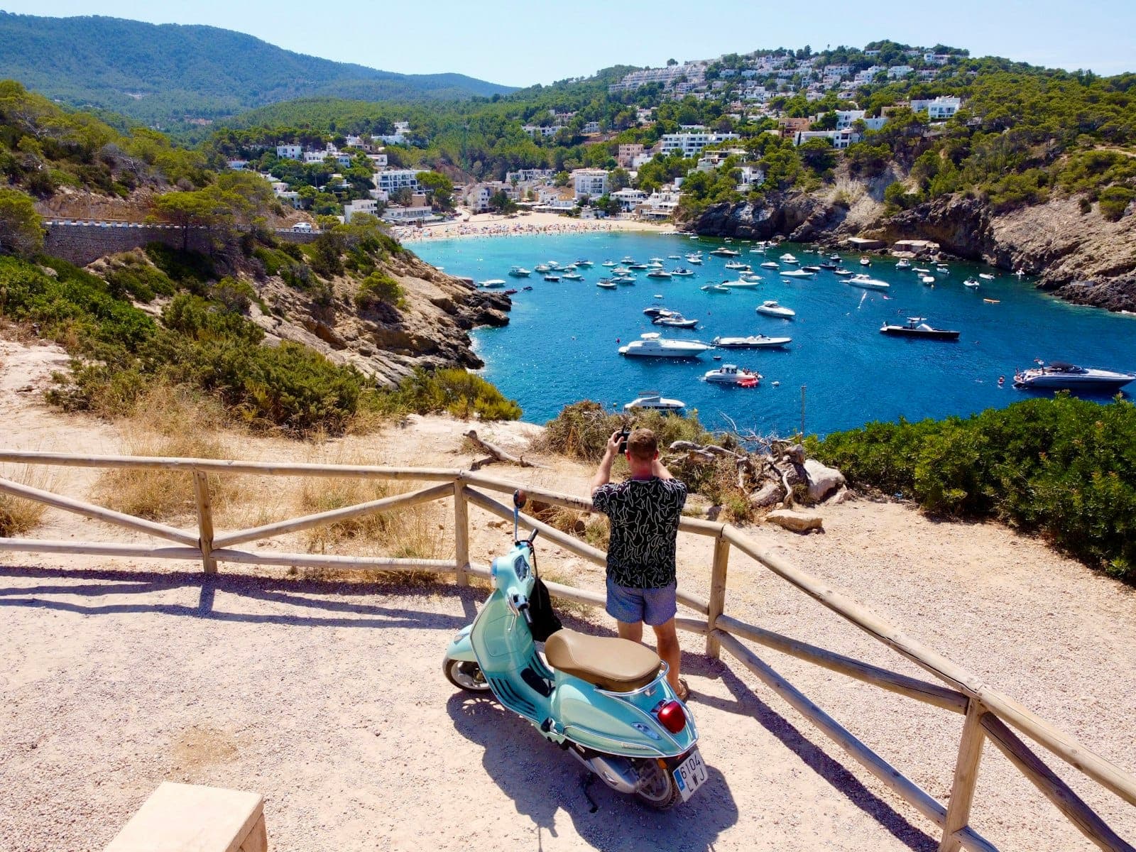 Person with a scooter overlooks a scenic Ibiza bay with boats, pine trees, hills, and a village in the background under a clear sky.
