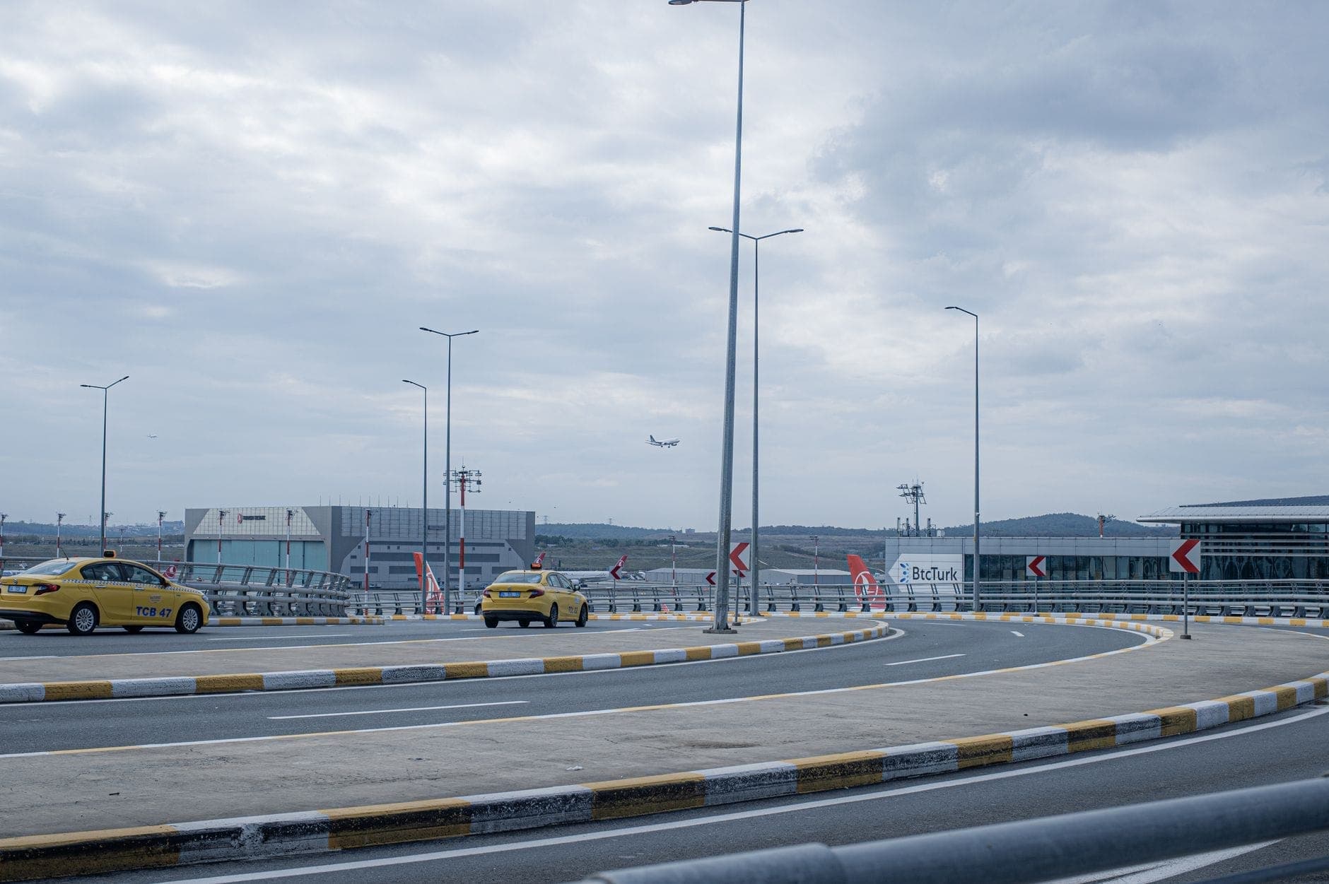 Estrada de acesso curva ao aeroporto com dois táxis amarelos, terminais do aeroporto e um avião decolando ao fundo sob um céu nublado