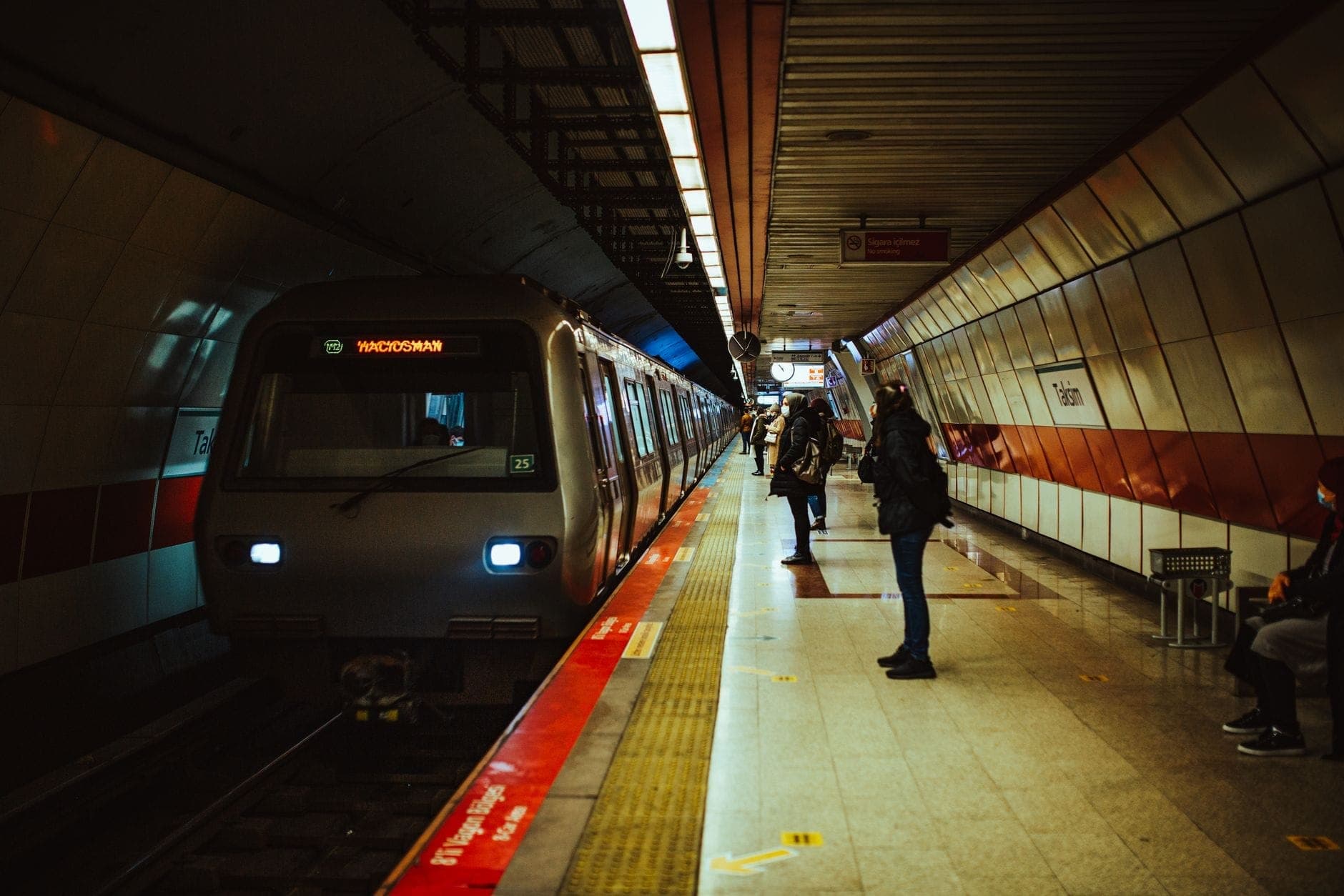 Estação de metrô de Istanbul com trem parado na plataforma, pessoas esperando e marcações de linha vermelha no chão.