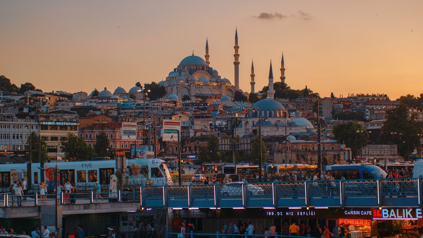 Istanbul bei Sonnenuntergang mit einem belebten Uferbereich – Fähren, eine Tram, Autos, Menschen und die historische Süleymaniye-Moschee im Hintergrund.