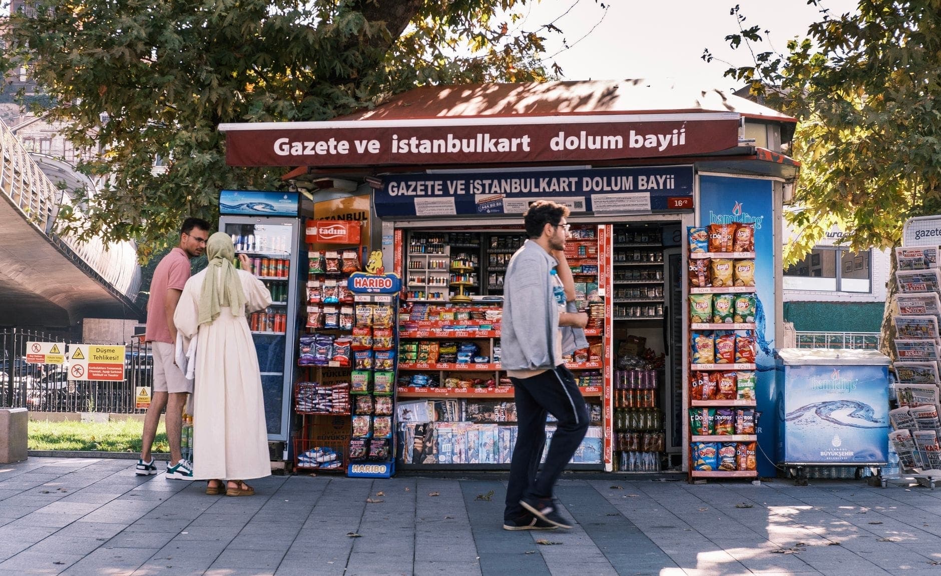 Quiosque de rua em Istambul com placa anunciando venda e recarga do Istanbulkart, pessoas comprando itens e passando pelo local.