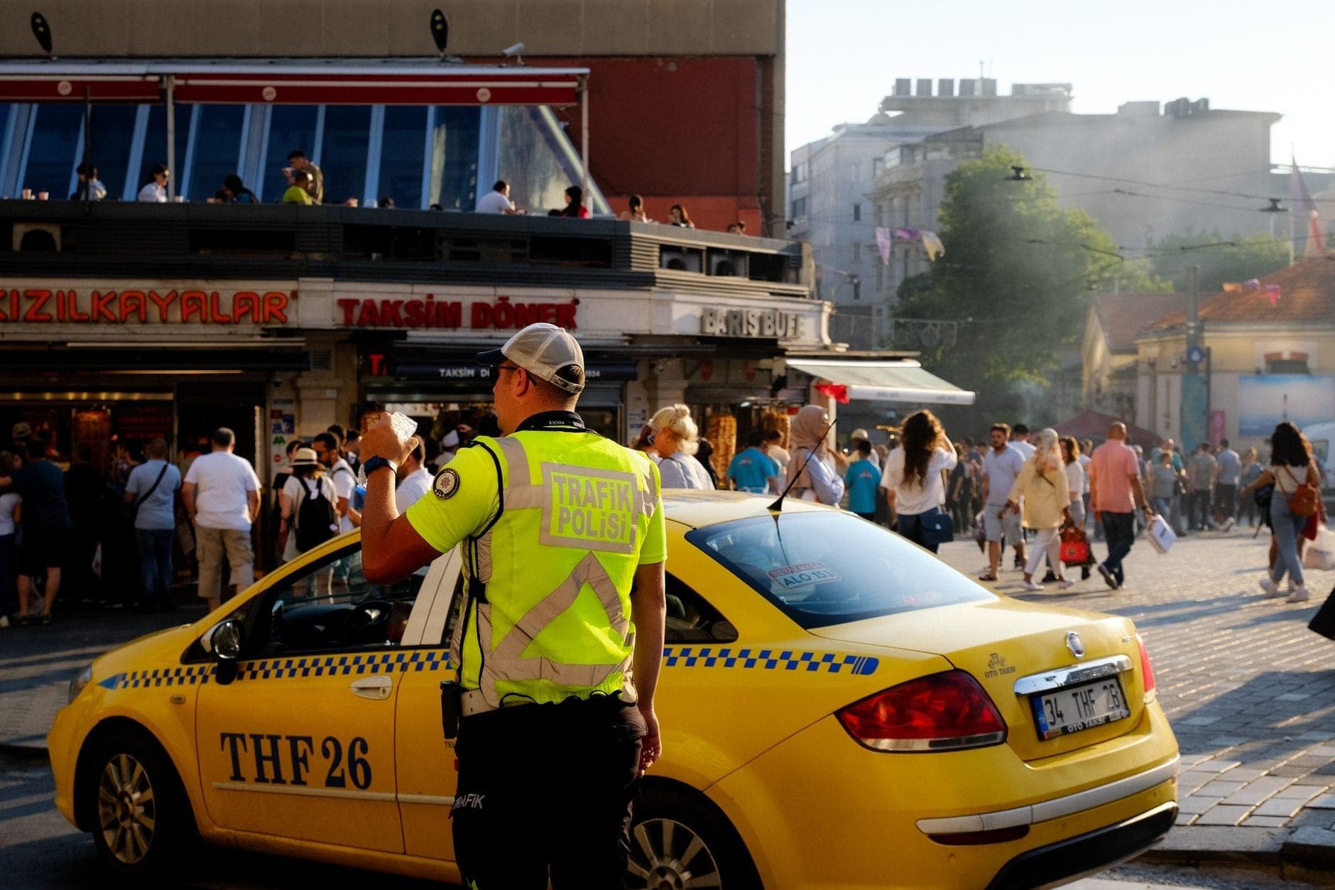 Táxi amarelo de Istambul parado em uma praça movimentada, com um agente de trânsito próximo e pessoas caminhando em frente a lojas com o letreiro Taksim Döner.