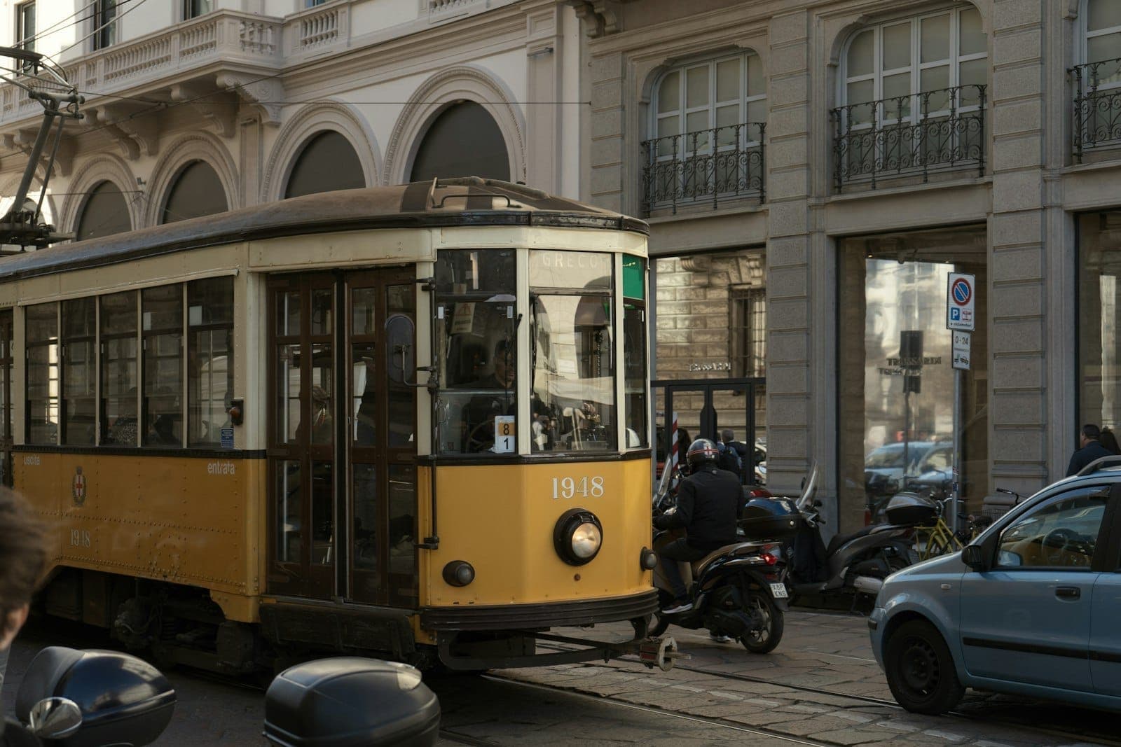 Classic yellow Milan tram traveling down a city street surrounded by historic buildings and parked scooters on a sunny day.