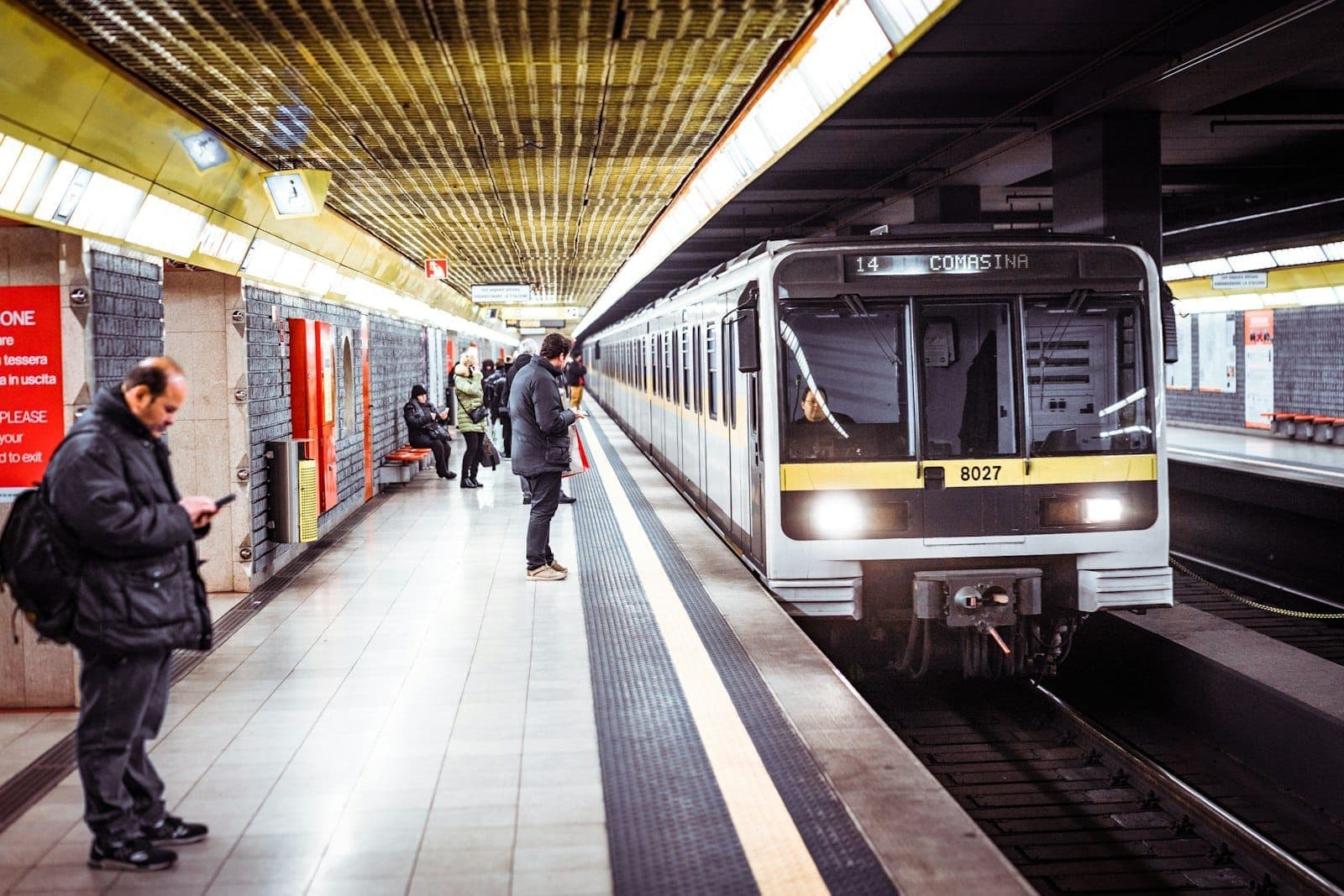 U-Bahn-Zug in Mailand fährt in einen unterirdischen Bahnsteig ein, auf dem Menschen warten und moderne gelbe Deckenleuchten leuchten.