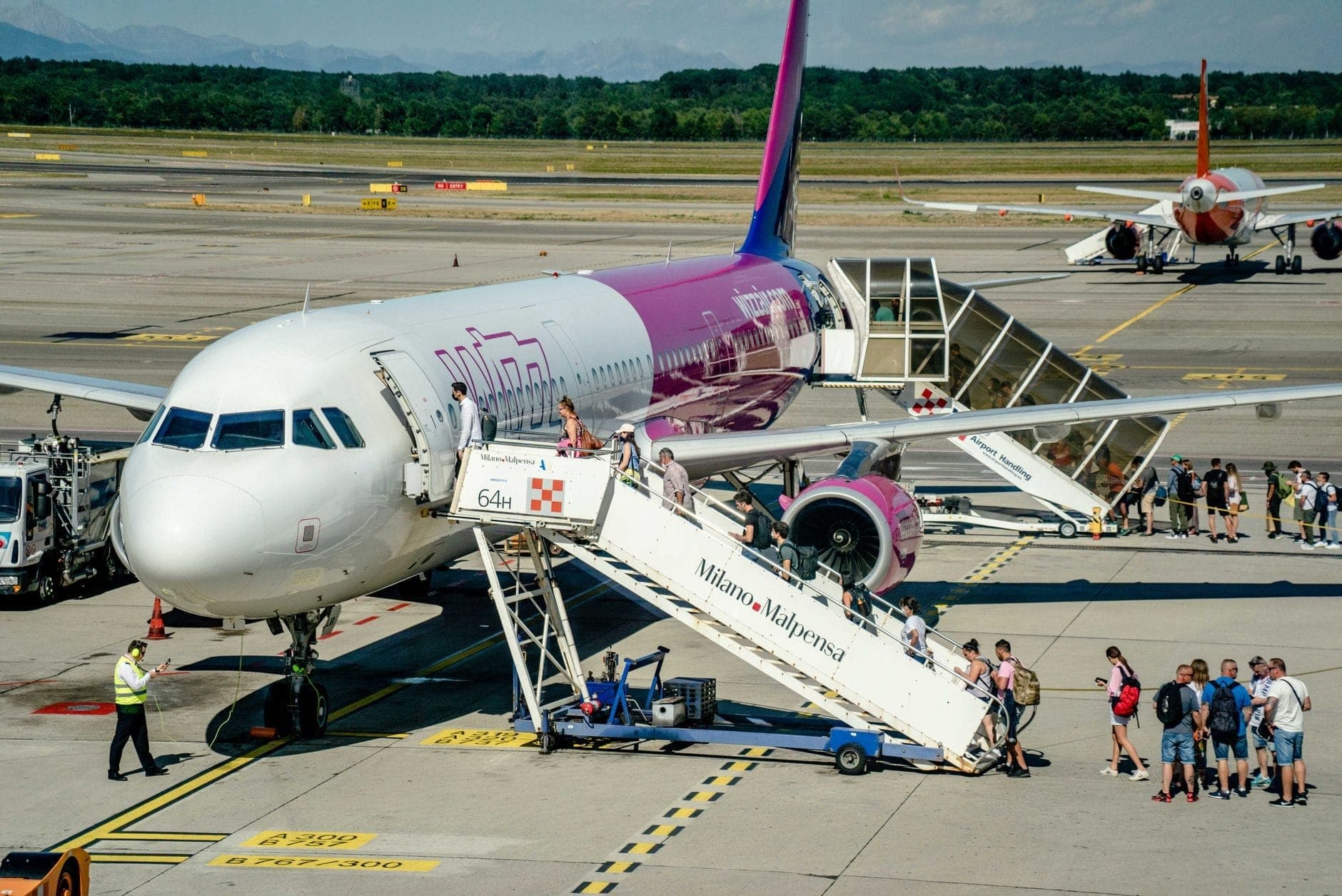 Passagiere, die über eine Treppe mit der Aufschrift 'Milano Malpensa' aus einem Flugzeug aussteigen, mit einem weiteren Flugzeug und einer Rollbahn im Hintergrund.