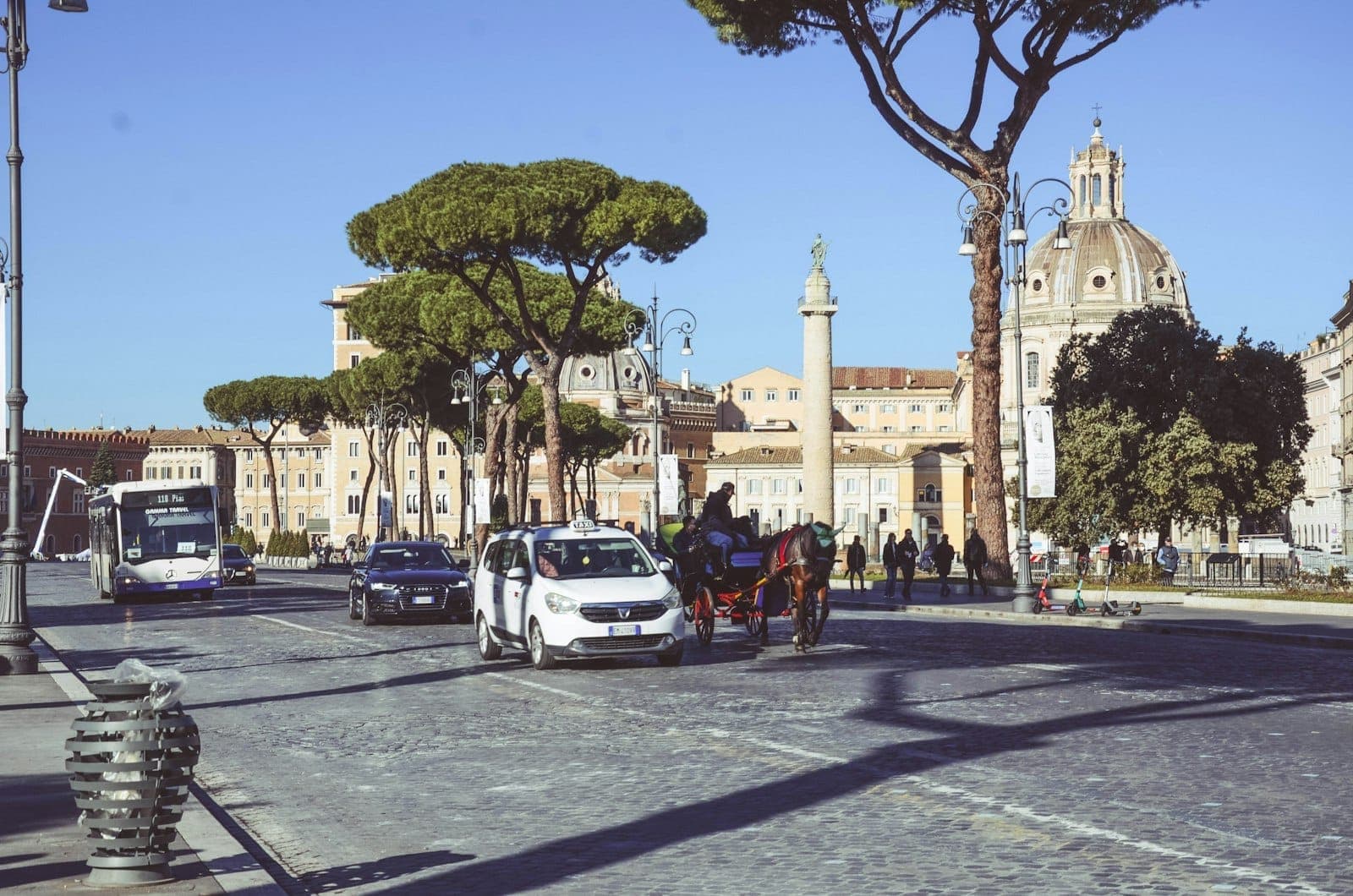 Đường phố rộng ở Rome với ô tô, taxi, xe buýt và xe ngựa, phía sau là các nhà thờ mái vòm và cây thông La Mã dưới bầu trời trong xanh.