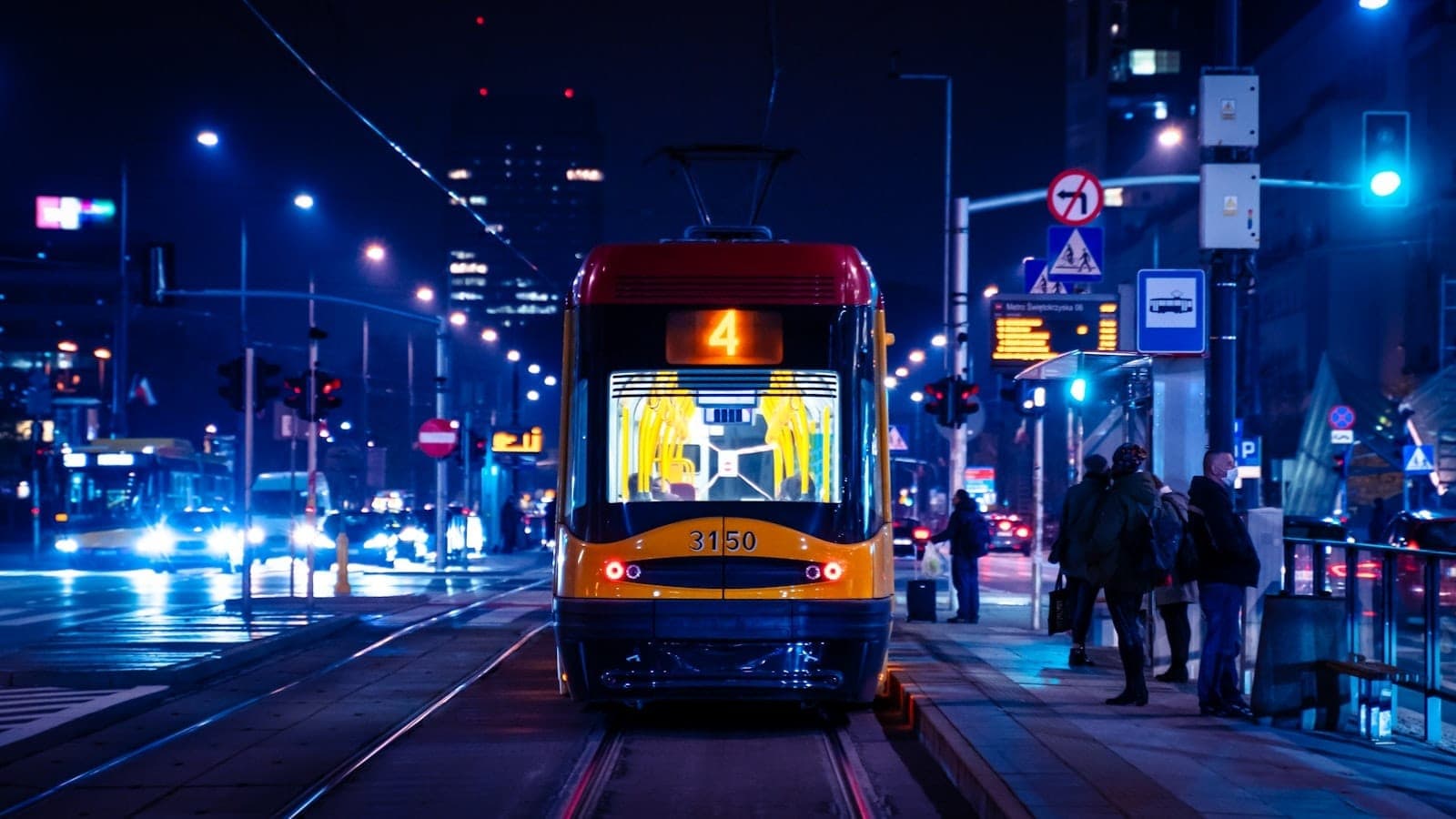 Moderne Warschauer Straßenbahn bei Nacht im Stadtzentrum, mit beleuchteten Straßenlaternen, wartenden Menschen an der Haltestelle und Stadtgebäuden im Hintergrund.