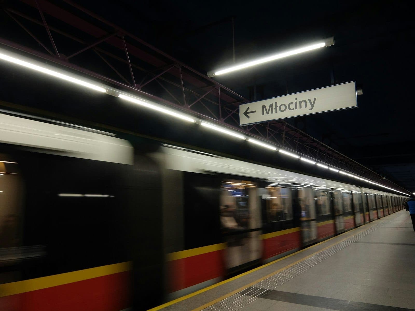 Peron metro Warsawa dengan kereta yang sedang bergerak dan tanda stasiun Mlociny di atas, menampilkan sistem transportasi kota modern.