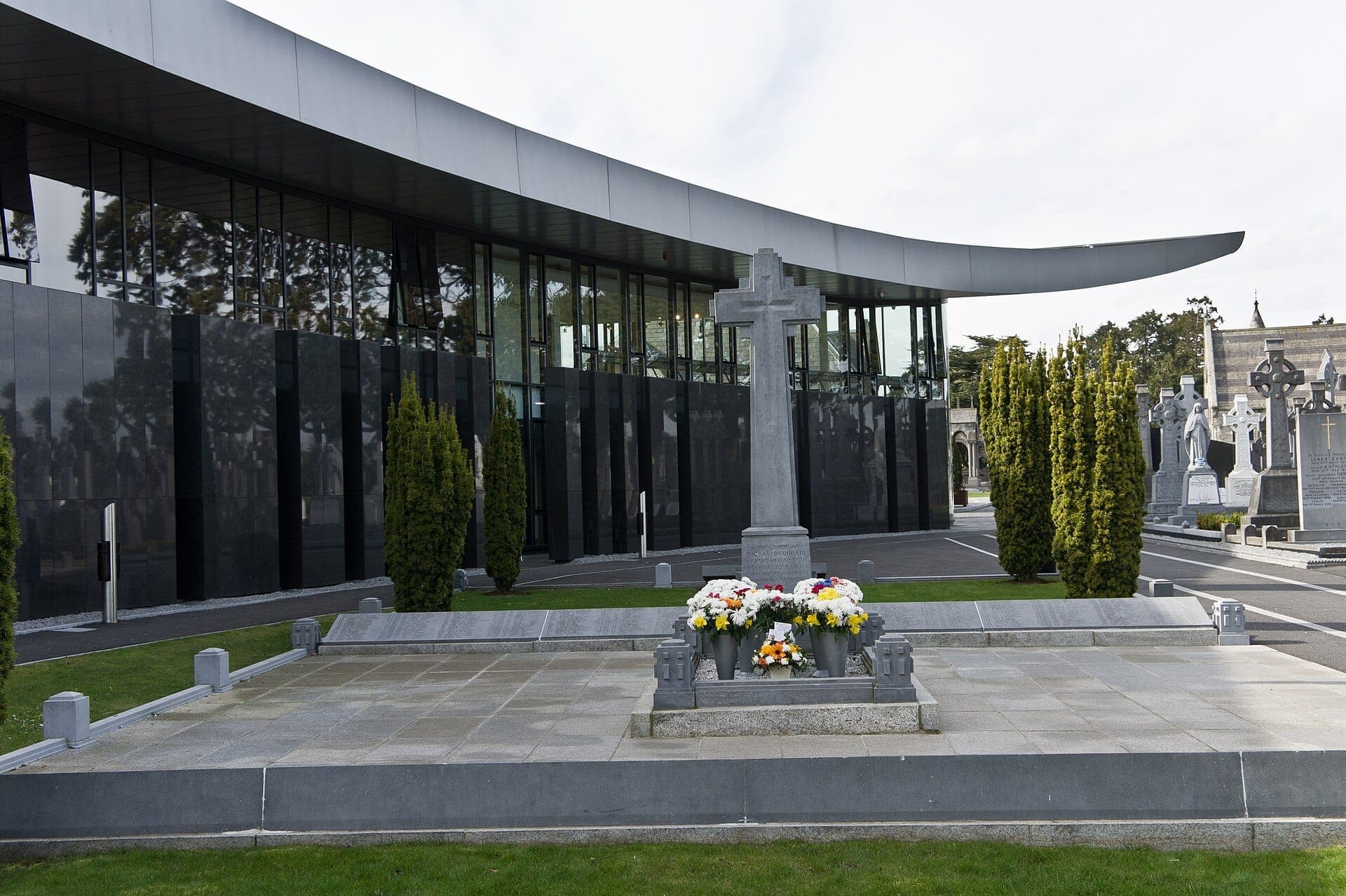 Nowoczesny szklany budynek Glasnevin Cemetery Museum z kamiennym monumentem w kształcie krzyża, kompozycjami kwiatowymi i zadbaną zielenią pod zachmurzonym niebem.