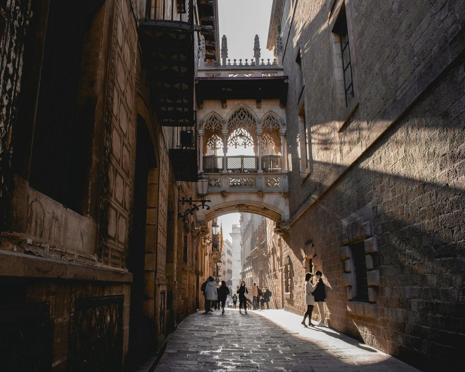 Isang makikipot na kalye ng cobblestone sa Gothic Quarter ng Barcelona na may mga sinaunang gusaling bato at ang tanyag na tulay na Pont del Bisbe, may sikat ng araw na sumasaklaw, at mga taong naglalakad.